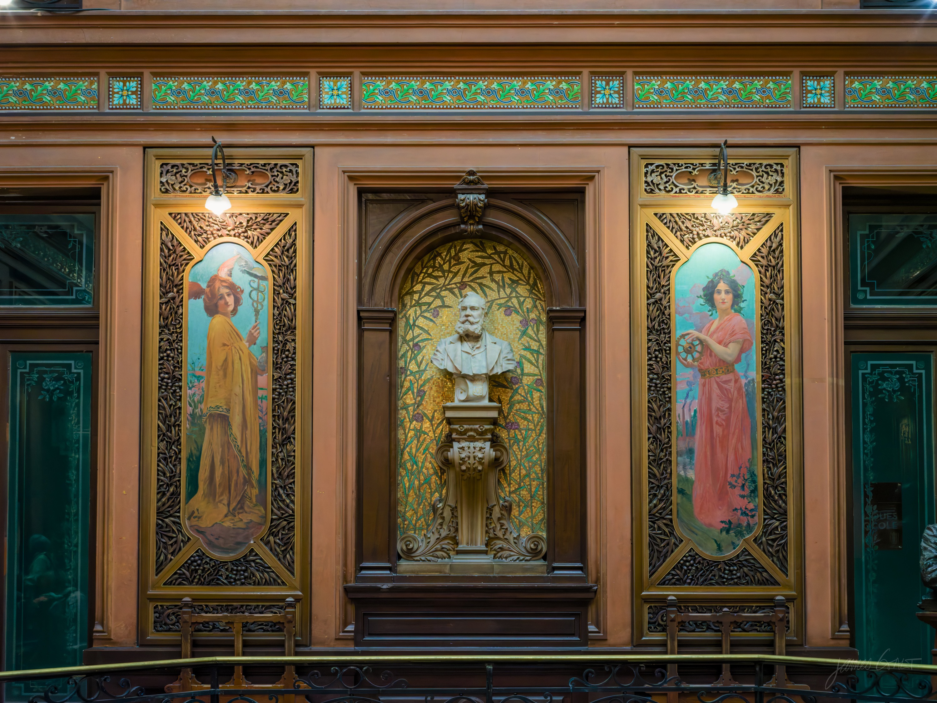 A carved wooden wall with a marble bust in a central niche flanked by two Art Nouveau painted panels of women, a green frieze running above.