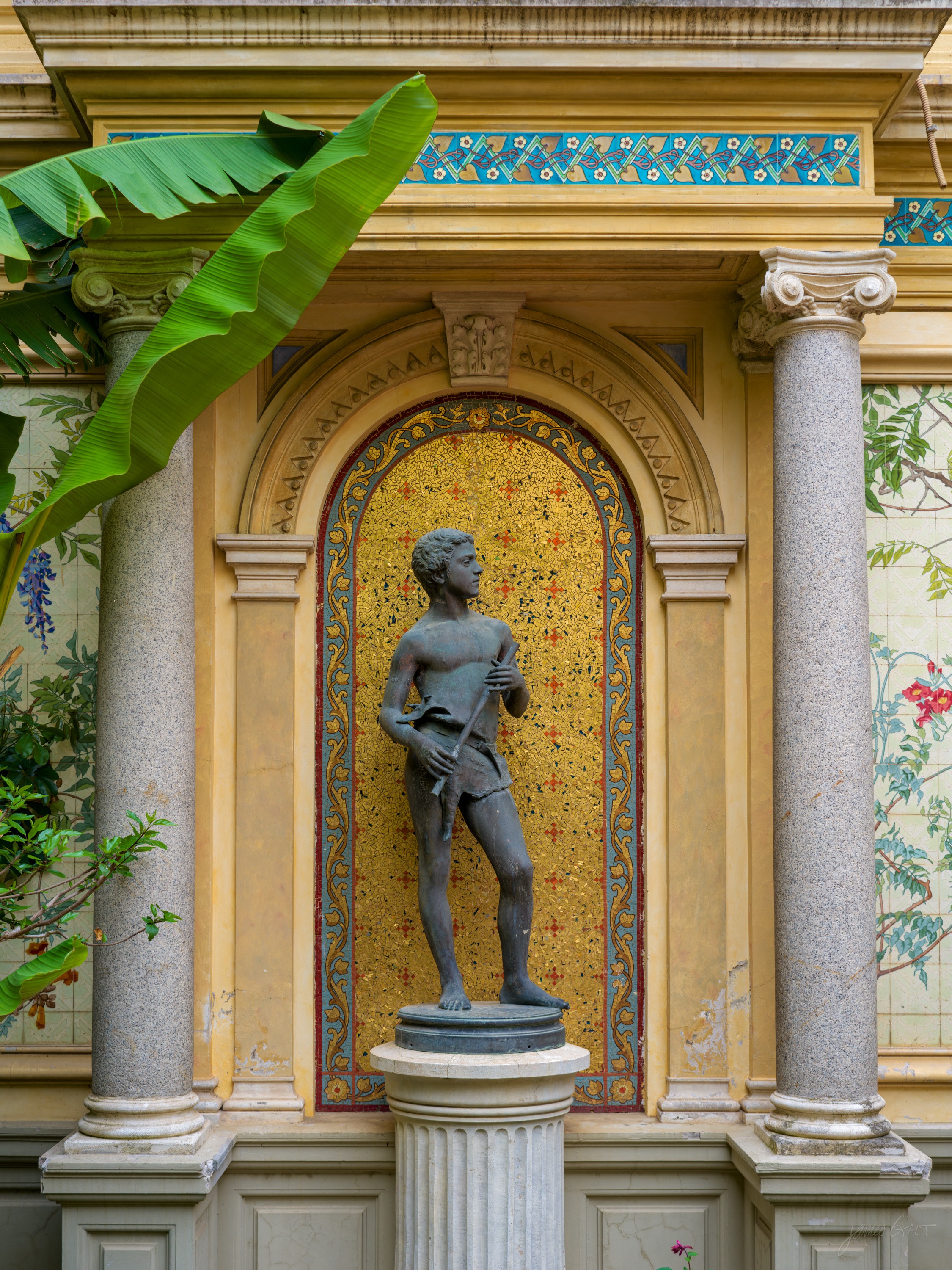 A bronze statue of a standing youth on a fluted plinth, set against a niche lined in gold mosaic between marbled columns, banana leaves reaching in from the left.
