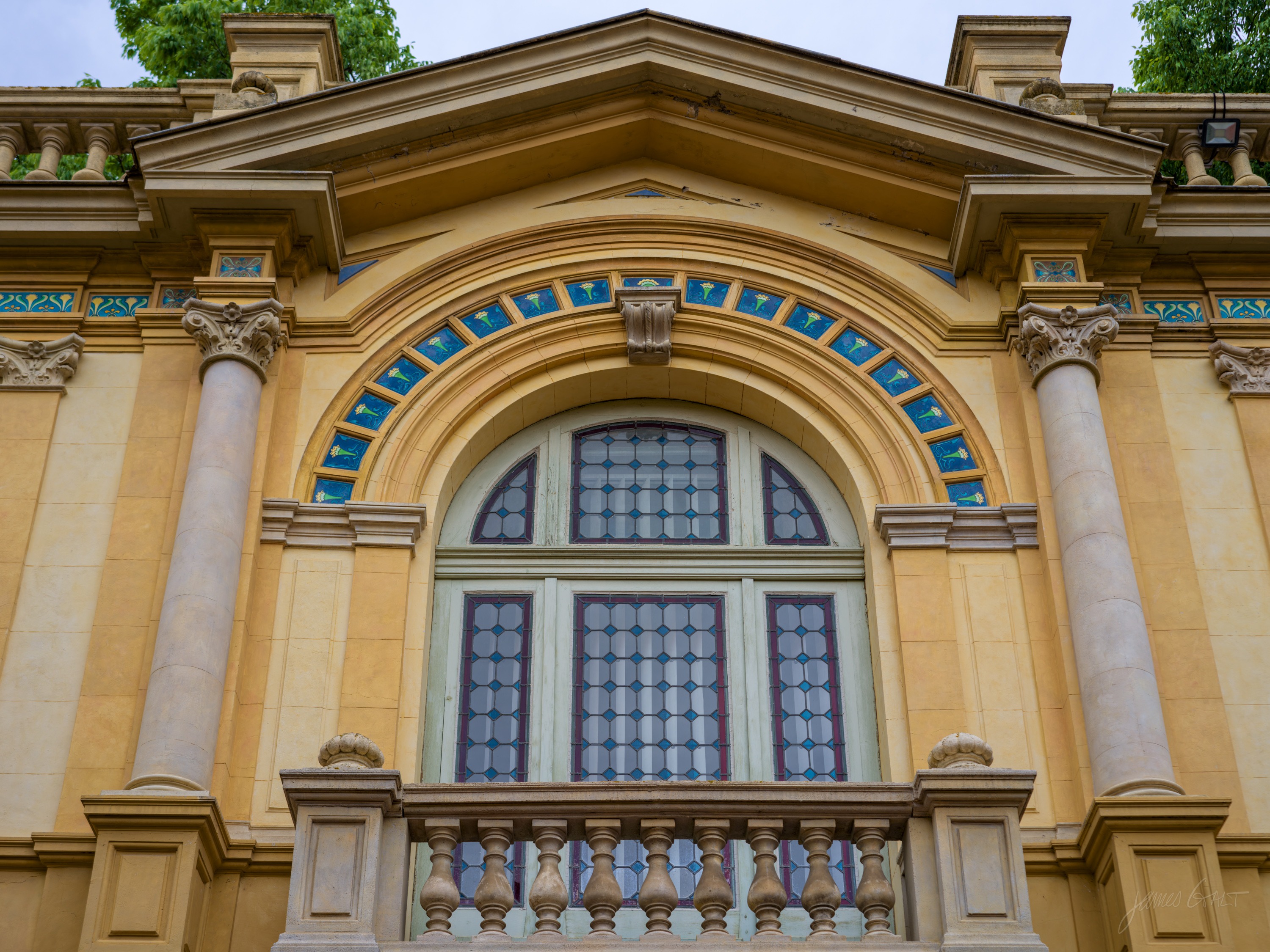 An ochre classical facade with a tall arched window under a broken pediment, paired columns and a stone balustrade below, blue-tiled detailing above.