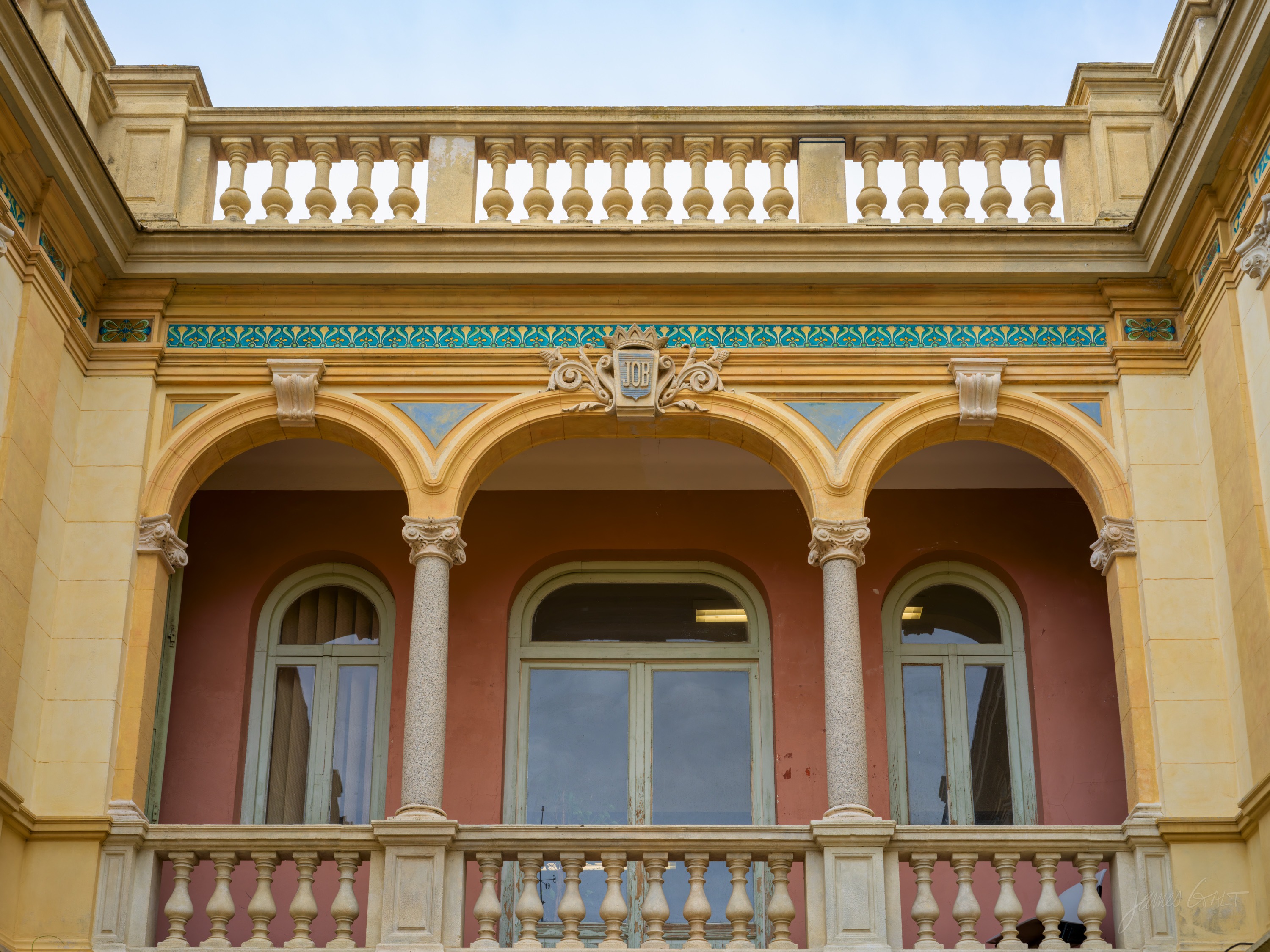 A first-floor loggia of round arches on slim pink columns above a balustrade, an ochre wall with a green-and-gold frieze, a crest set at the centre.