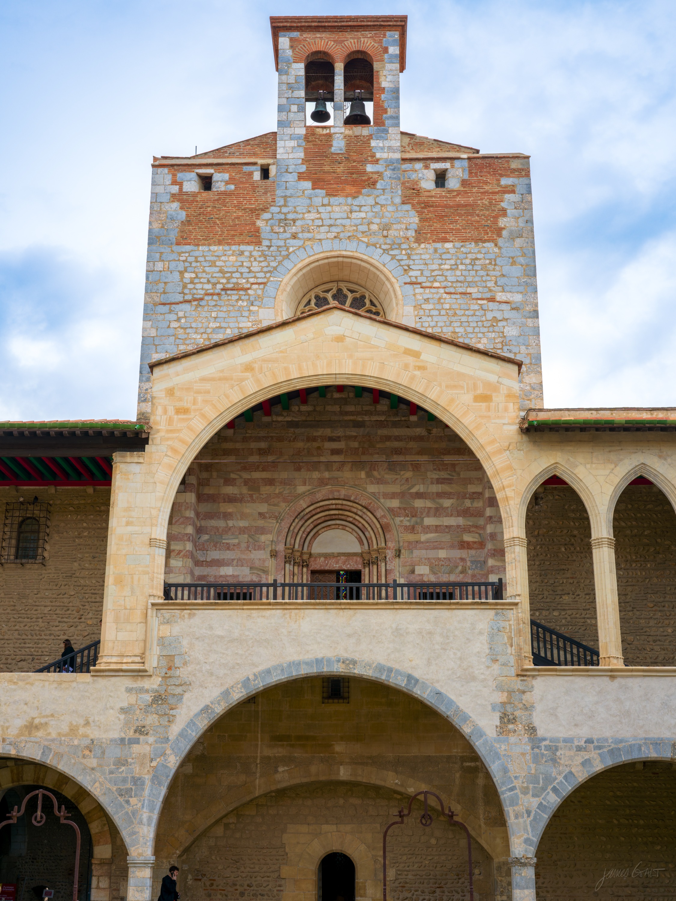 A two-storey stone courtyard facade with a bell tower above, a round-arched open loggia and a smaller arcaded gallery, under grey cloud.