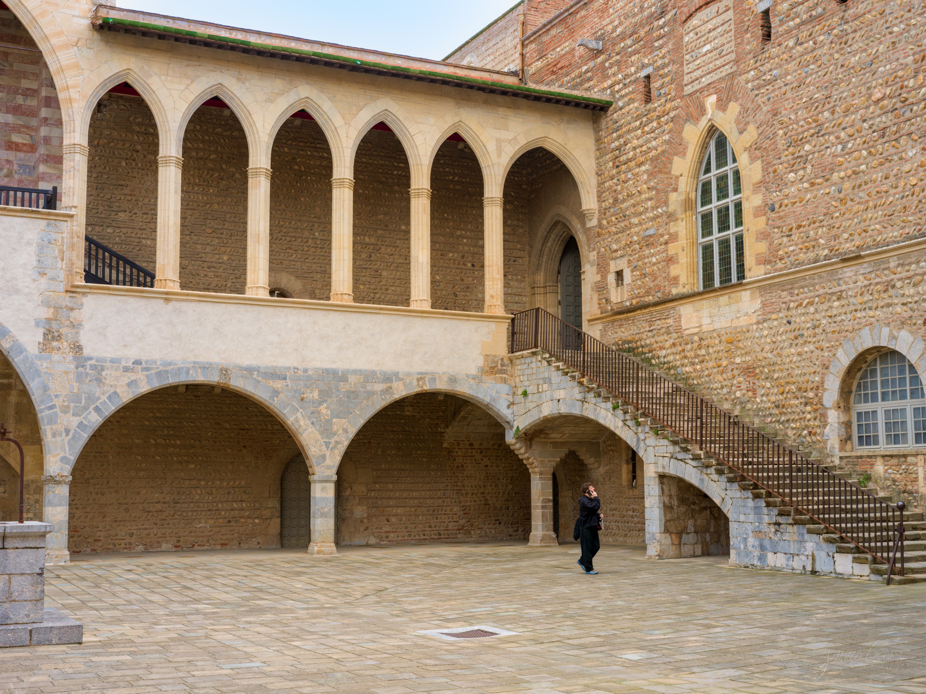 A galleried courtyard with a pointed-arch stone loggia above and vaulted arcades below, a broad stair climbing one side, a lone figure crossing the paving.