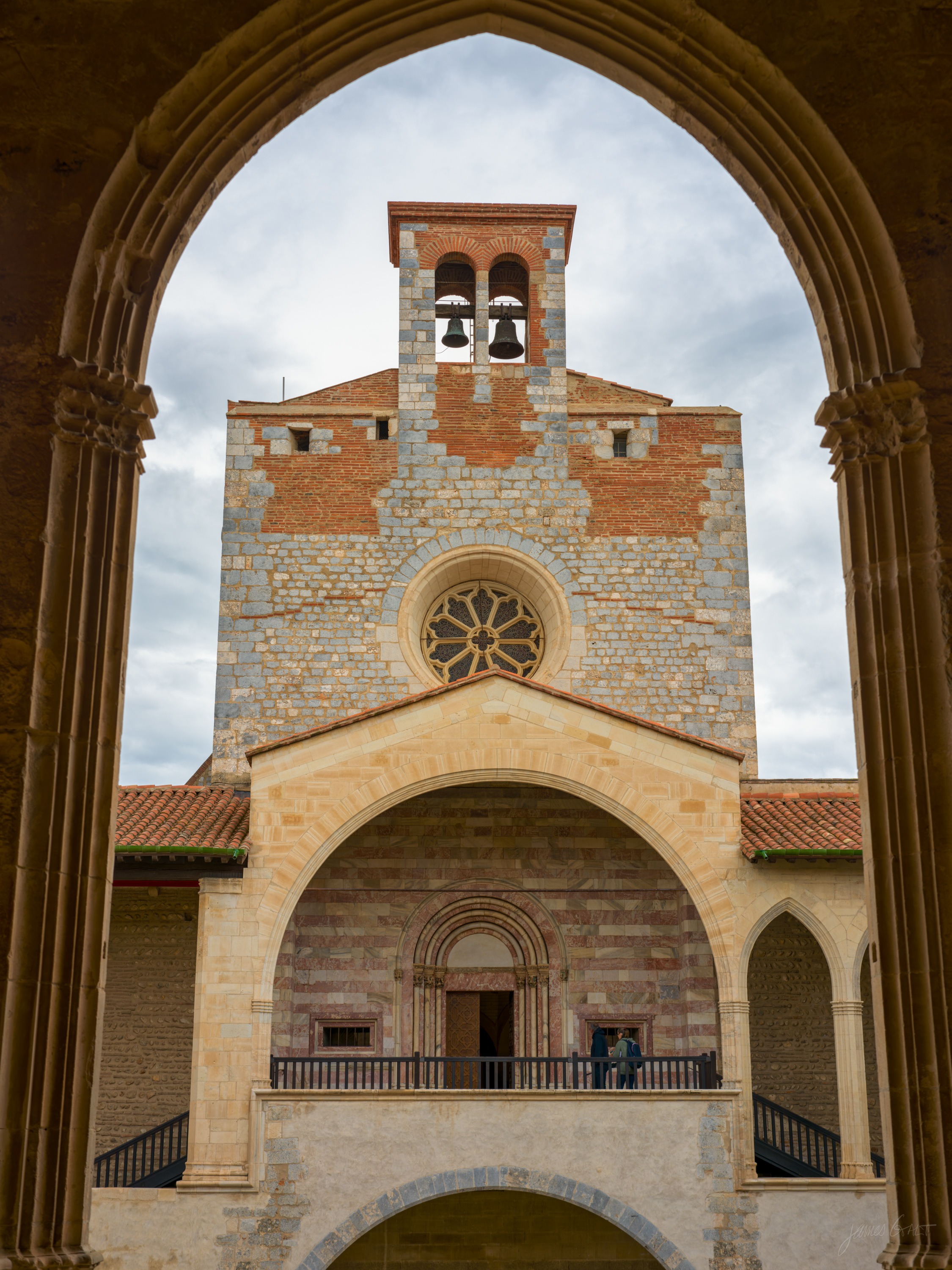 A stone chapel front with a bell gable and rose window, seen framed through a tall pointed Gothic arch in the foreground.