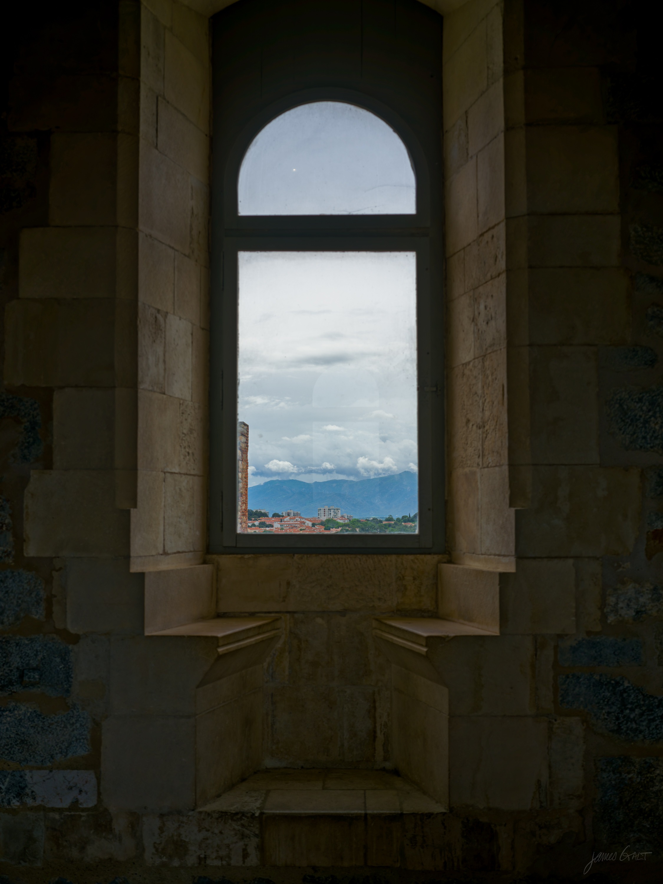 A deep stone window embrasure with a tall arched window, framing a distant snow-streaked mountain and a town on the plain under cloud.