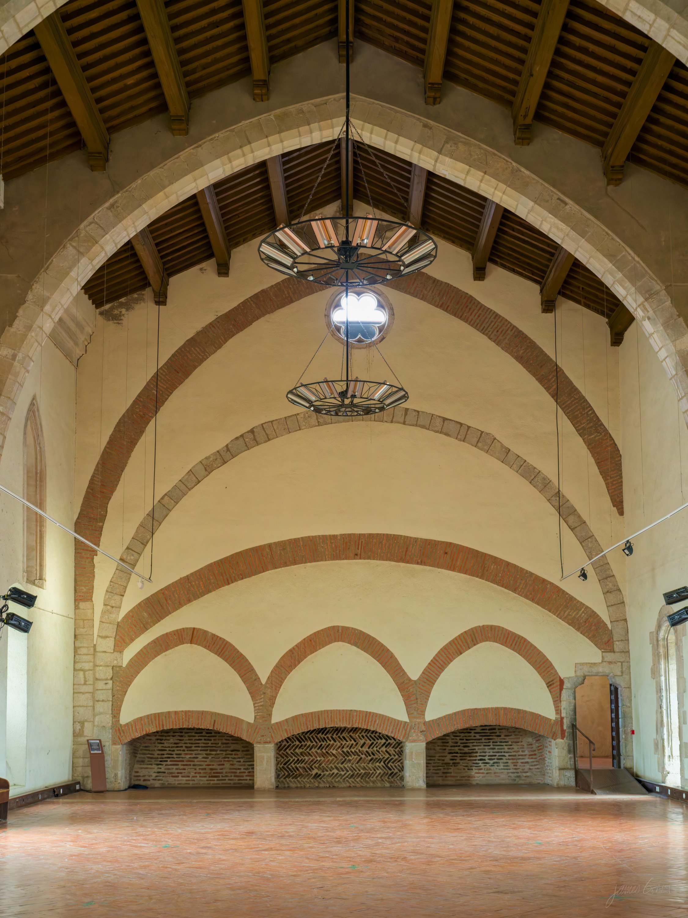 A vast empty hall with a timber roof and a wall of stepped blind brick arches at the far end, two iron lanterns hanging, warm light on the floor.