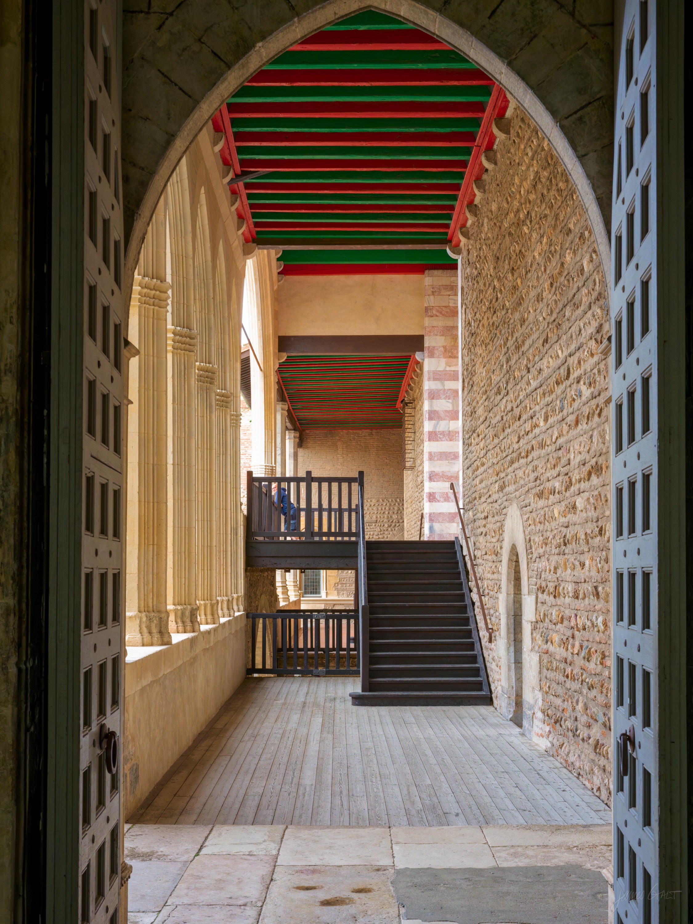 An open loggia with a boldly painted red and green timber ceiling, tall stone piers, and a dark staircase climbing beyond a heavy door.