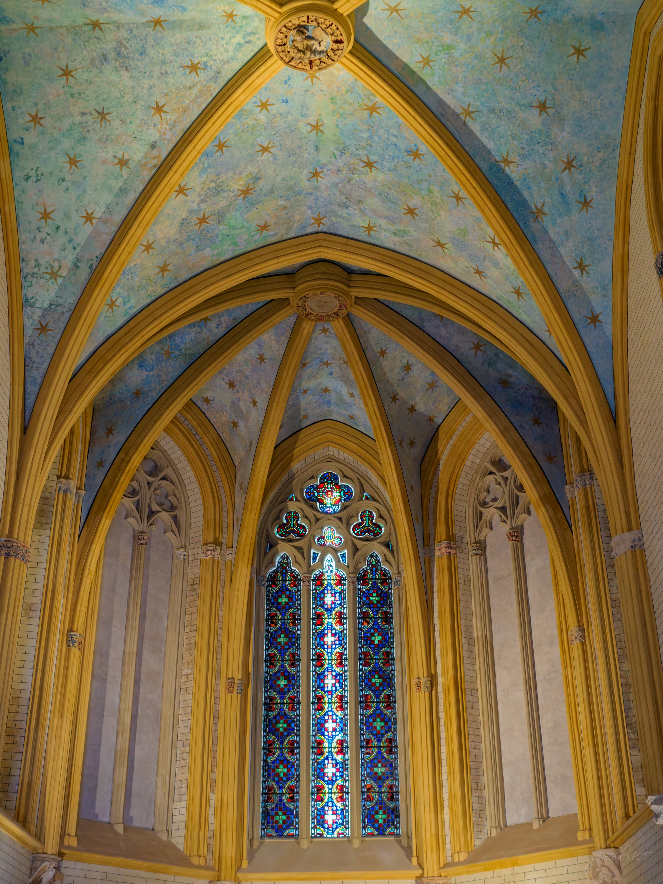 A Gothic chapel vault painted deep blue and scattered with gold stars, gilded ribs descending to a tall stained-glass window over the altar.