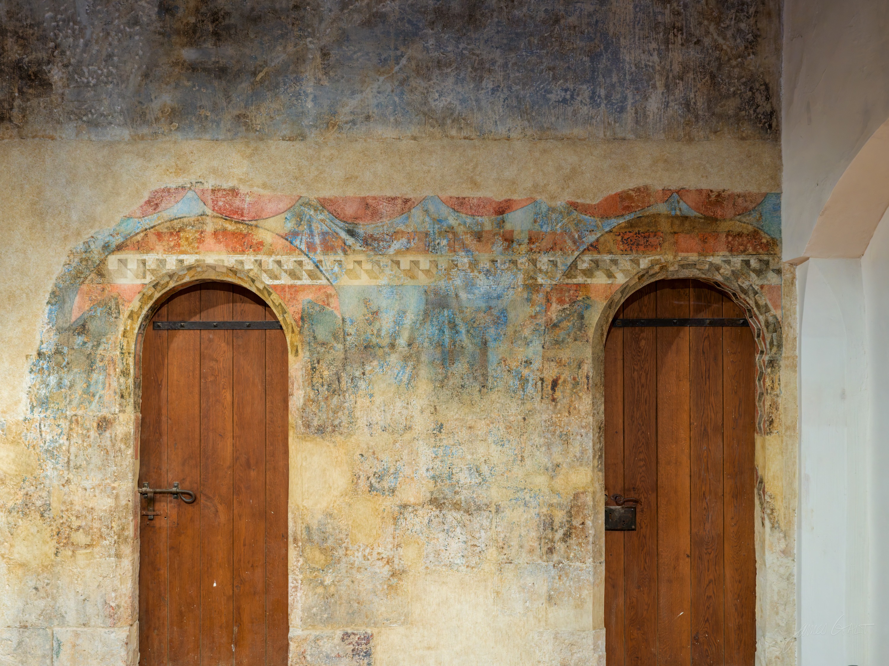 A rough plaster wall carrying two arched wooden doors, a band of faded red and blue medieval painting surviving above them.
