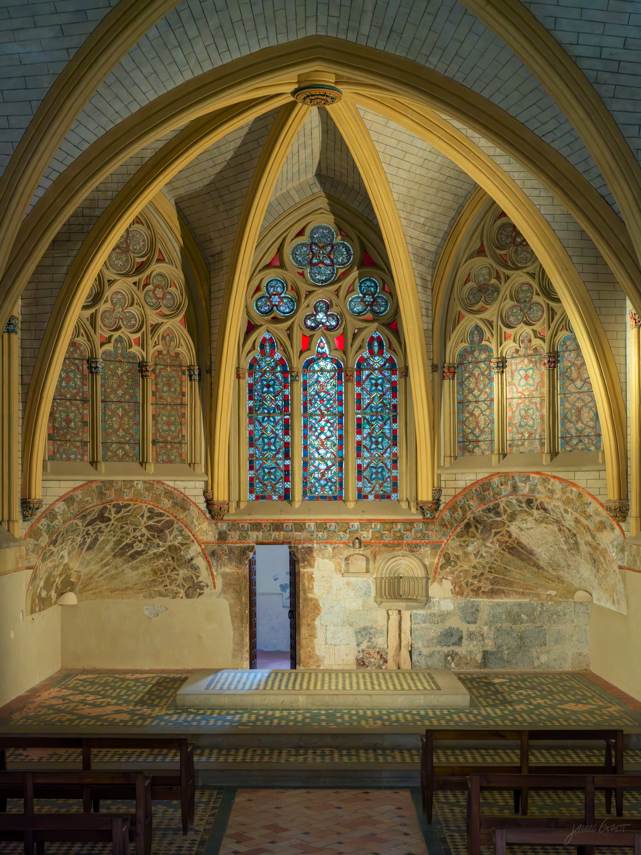 A vaulted chapel apse ringed with stained-glass windows above walls of faded painted decoration, empty benches on a tiled floor below.