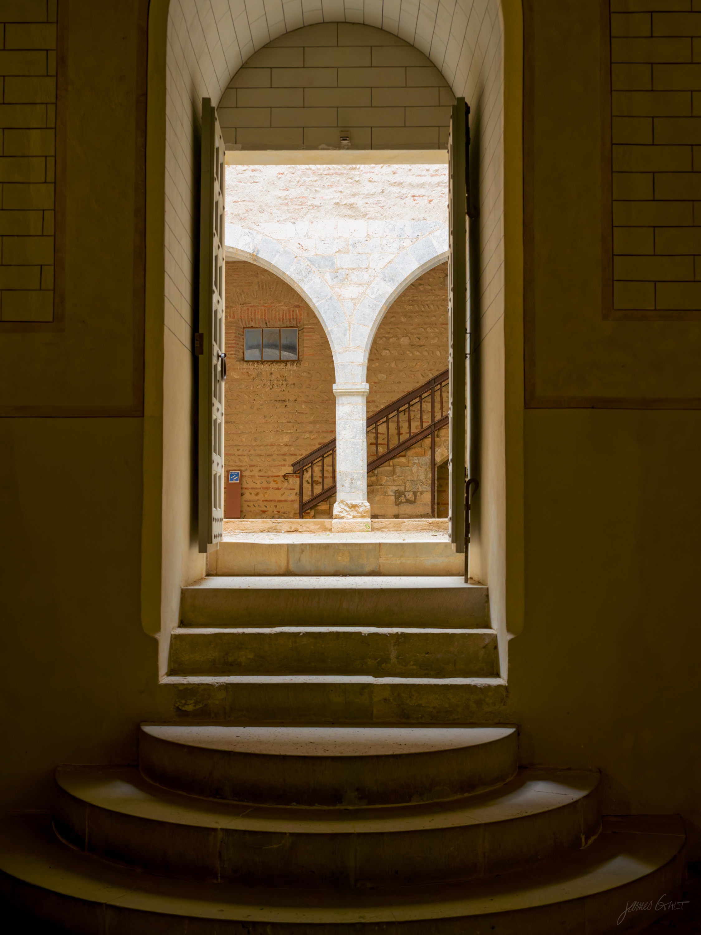 A stone stairwell arch framing a bright courtyard beyond, a single column and a flight of steps visible through the opening.