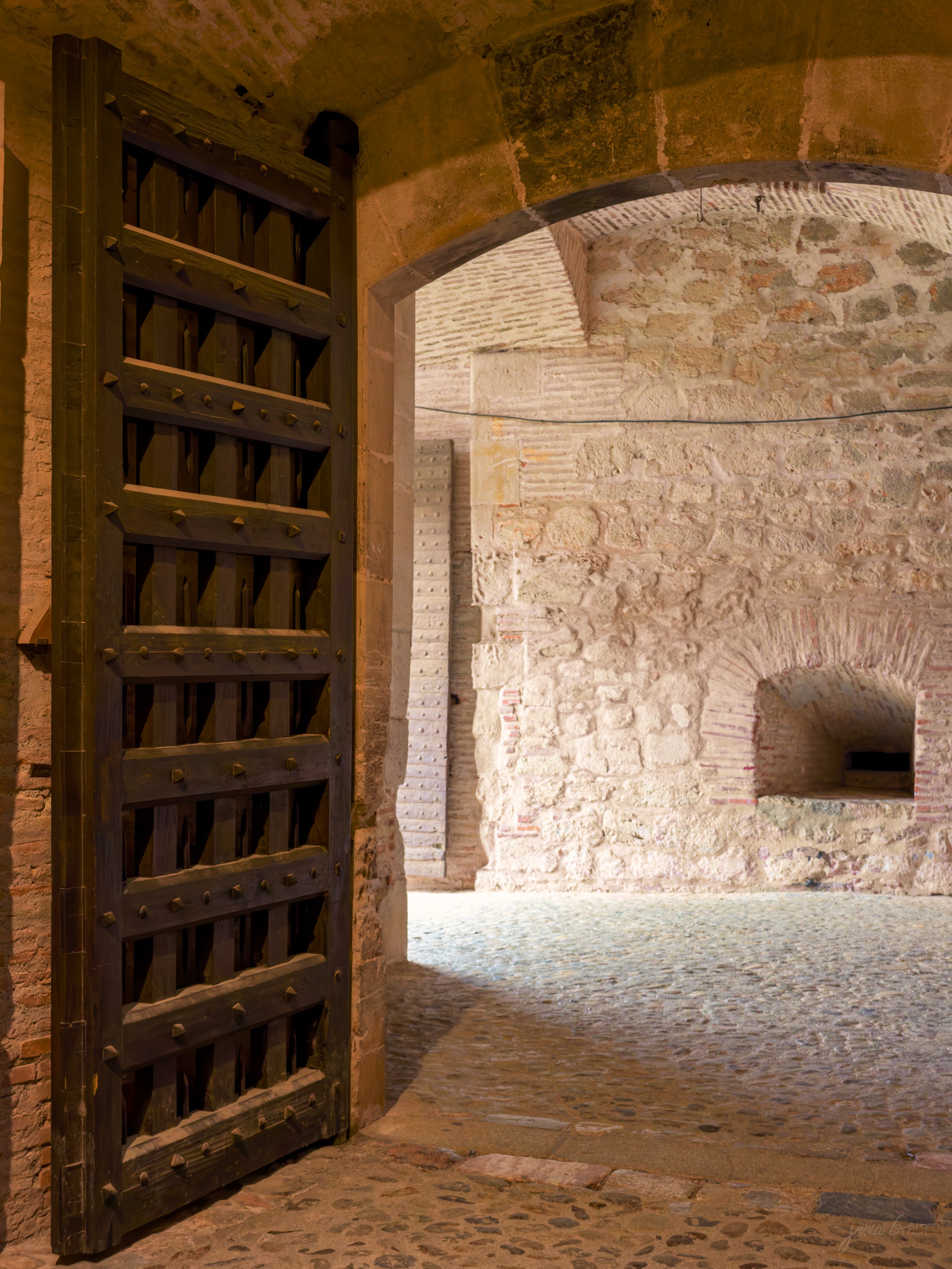 A dim vaulted interior with a heavy studded wooden door standing open, a domed brick oven niche set into the rough stone wall beyond.