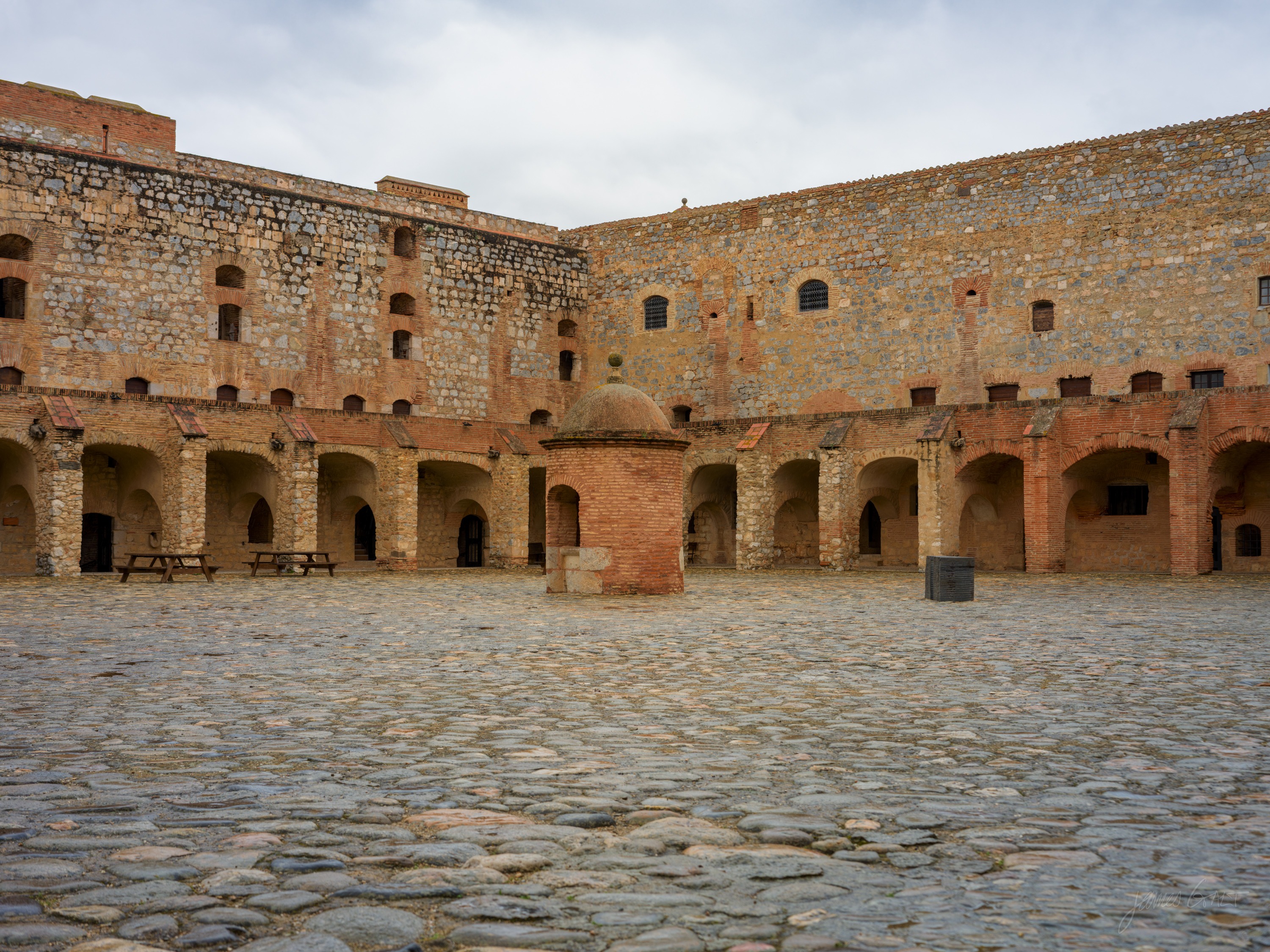 A cobbled fortress courtyard ringed by arched brick and stone galleries, a small domed well head at its centre under a pale sky.