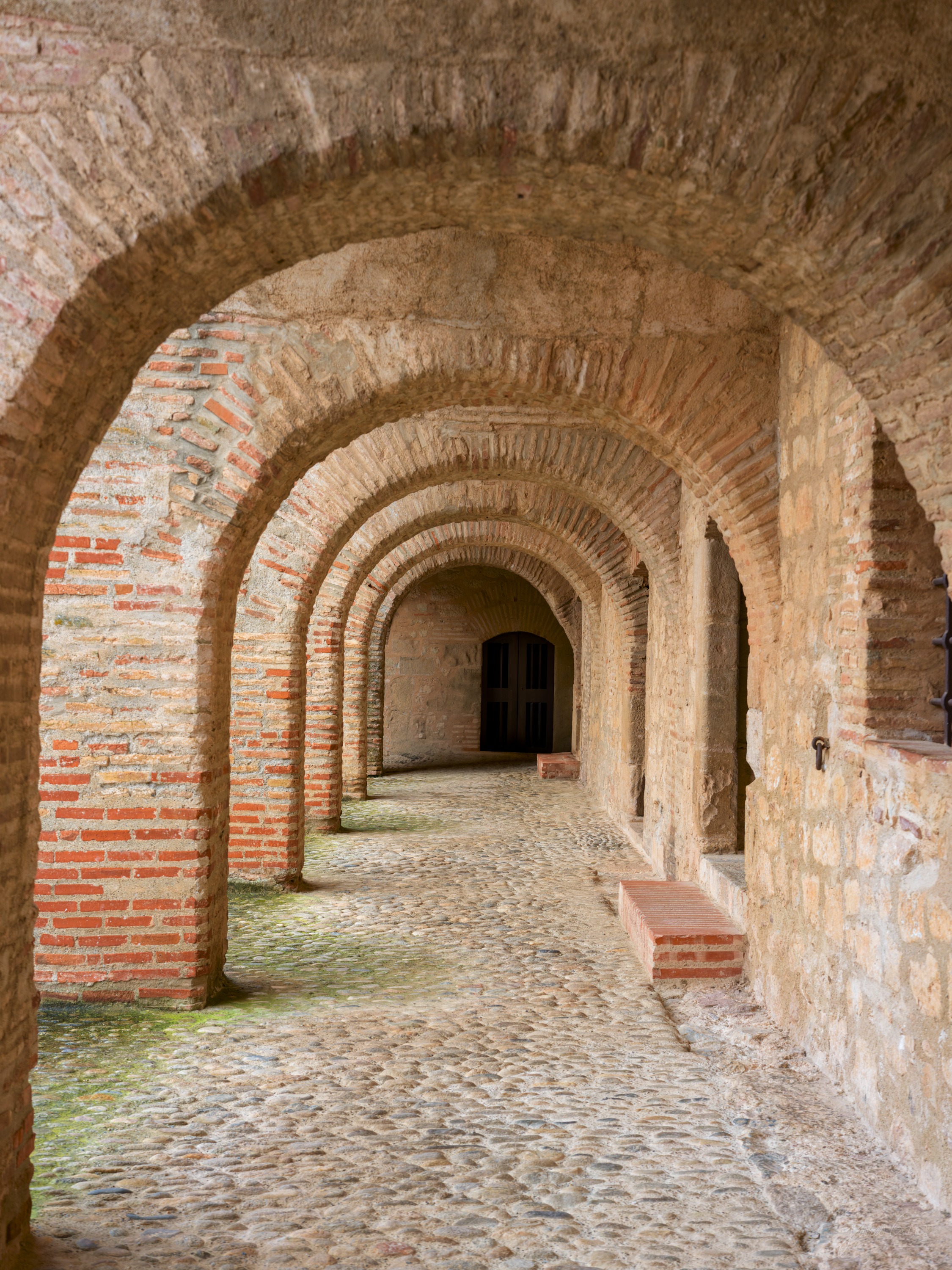 A long brick arcade of repeating arches over a cobbled floor, receding to a dark doorway, stone piers with brick voussoirs down one side.