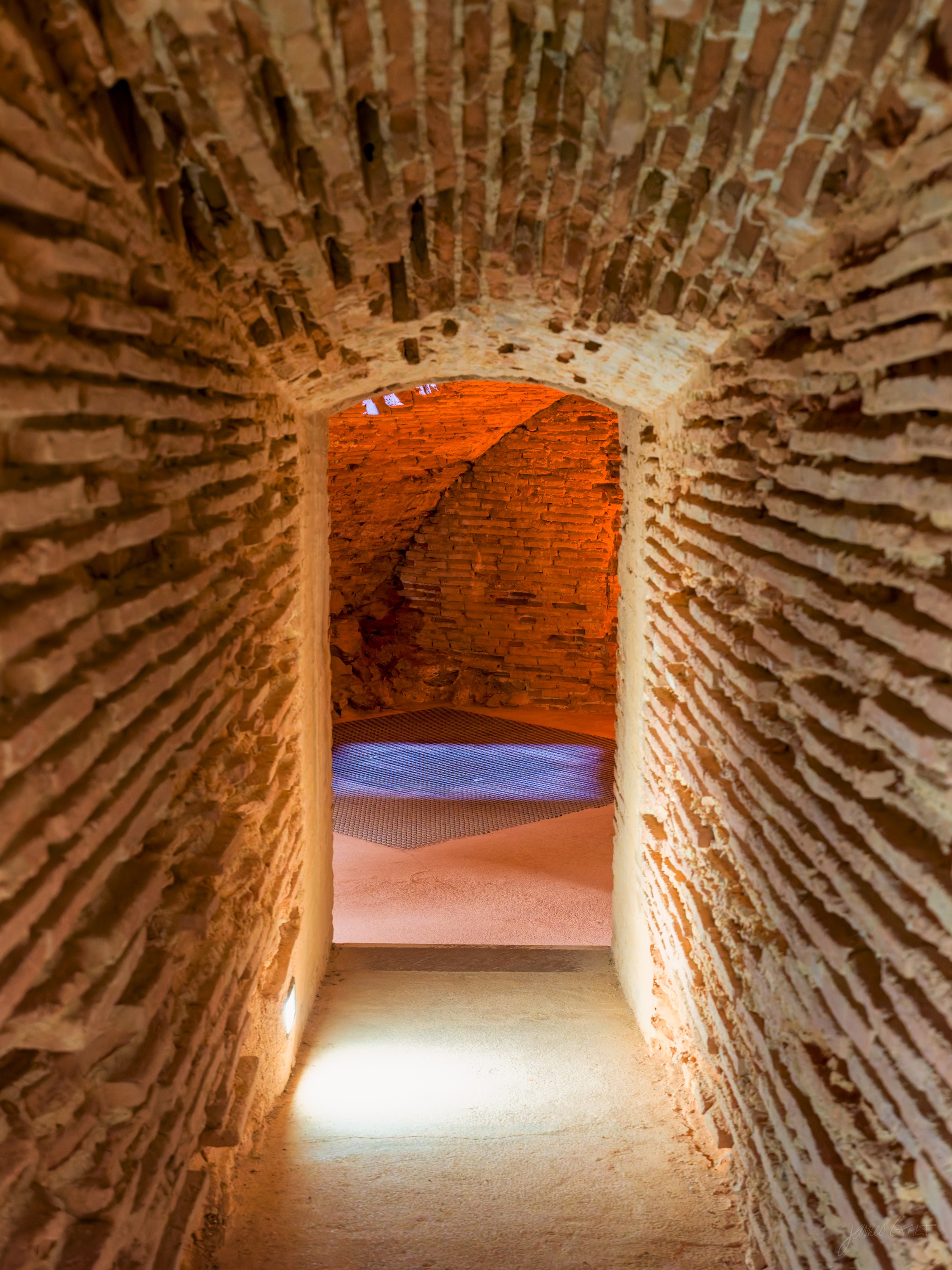 A low brick tunnel curving through thick walls, a shaft of daylight falling on the floor from an opening at the far end, otherwise near dark.