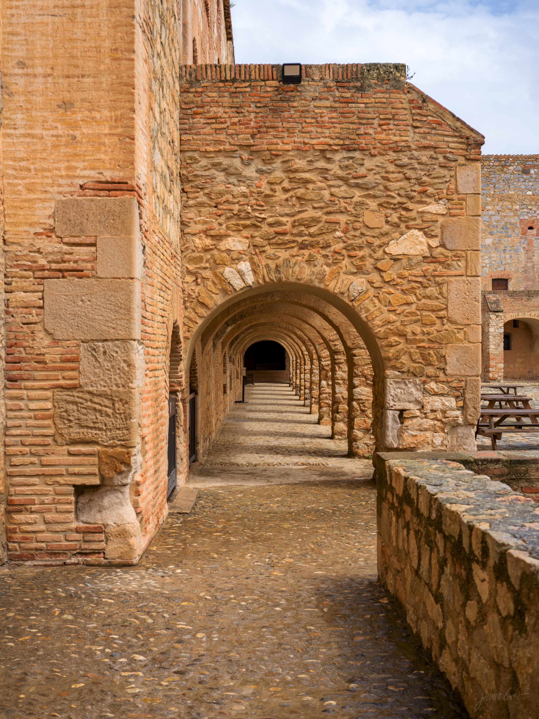 An open arcaded passage along a fortress wall, brick arches on one side and a low parapet on the other, cobbles running into the distance.