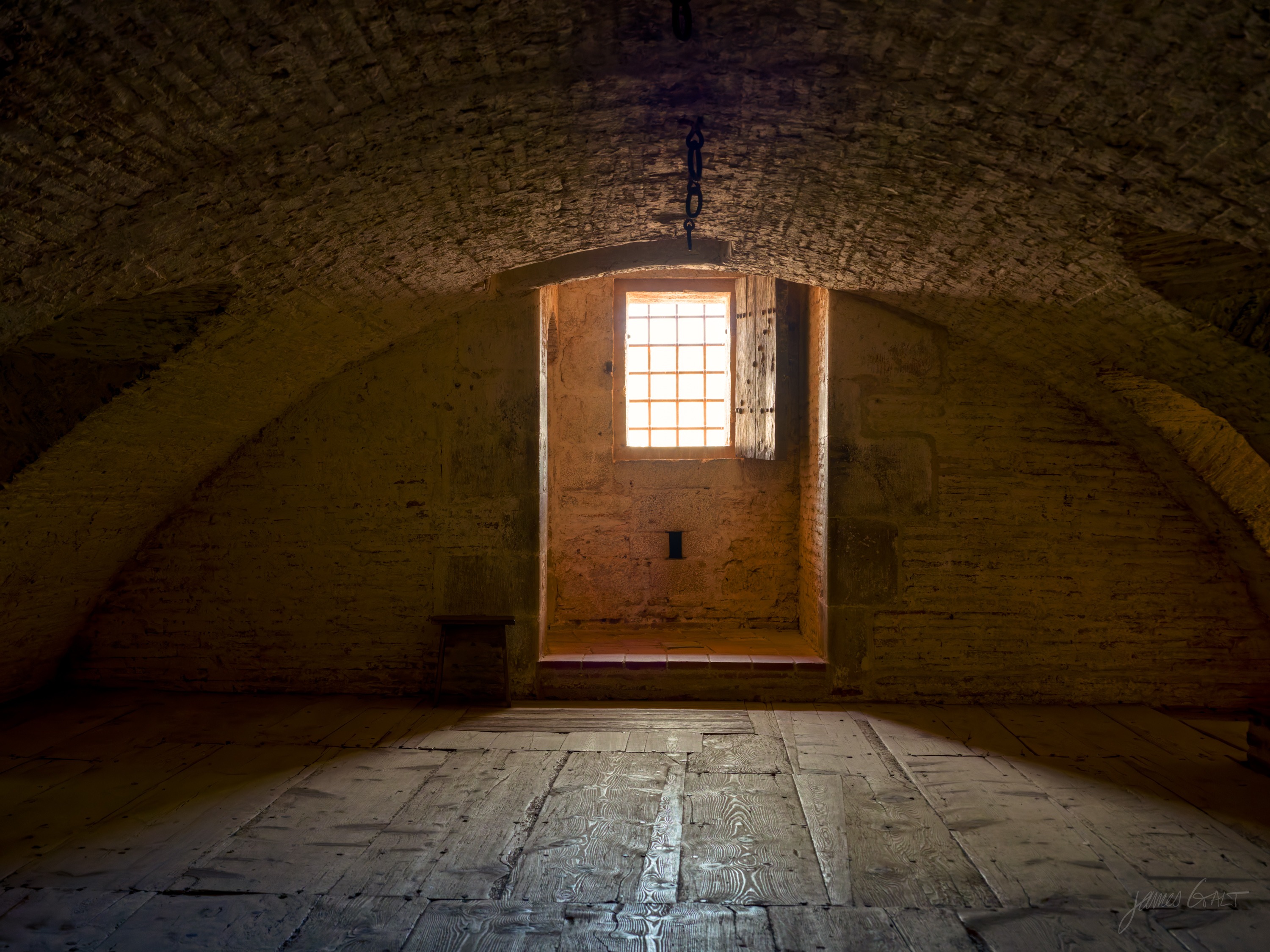 A broad brick barrel-vaulted room, mostly in shadow, a single barred window in the end wall throwing warm light across a worn plank floor.