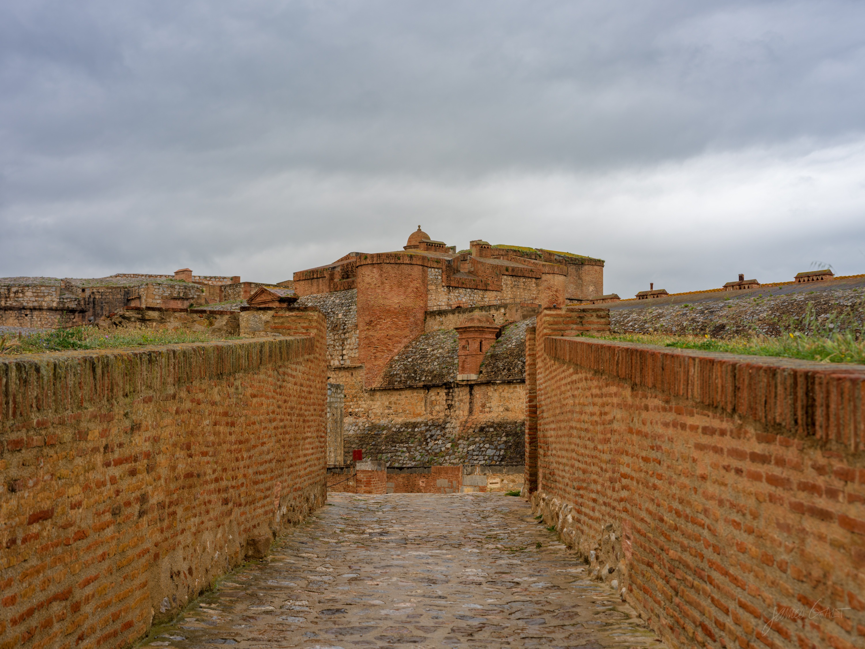 A cobbled causeway between high pink-brick and stone ramparts leading toward the squat towers of a fortress under a grey sky.