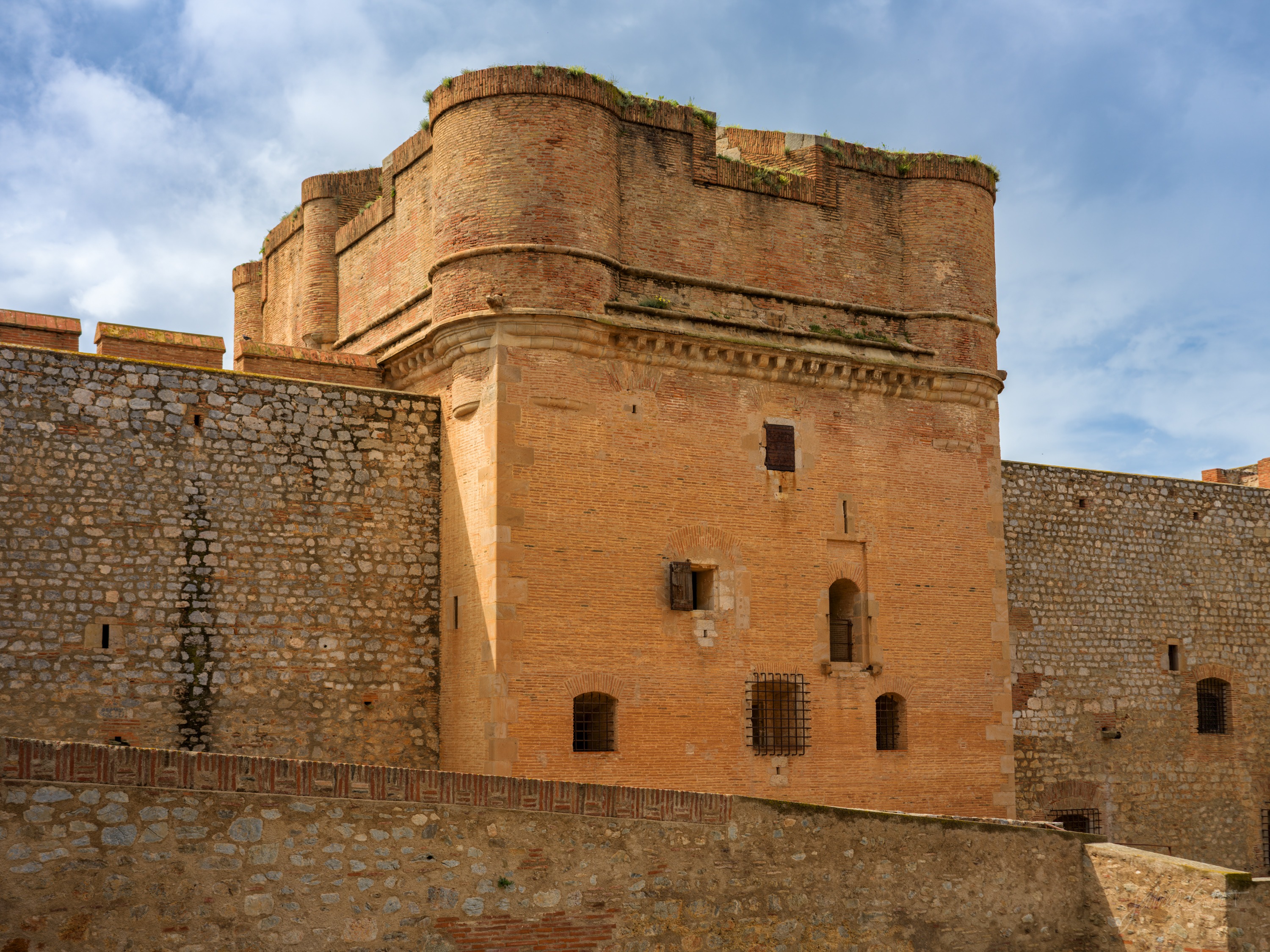 A massive rounded keep of pink brick over a stone base, small windows and gun ports in its face, set against a blue sky with cloud.