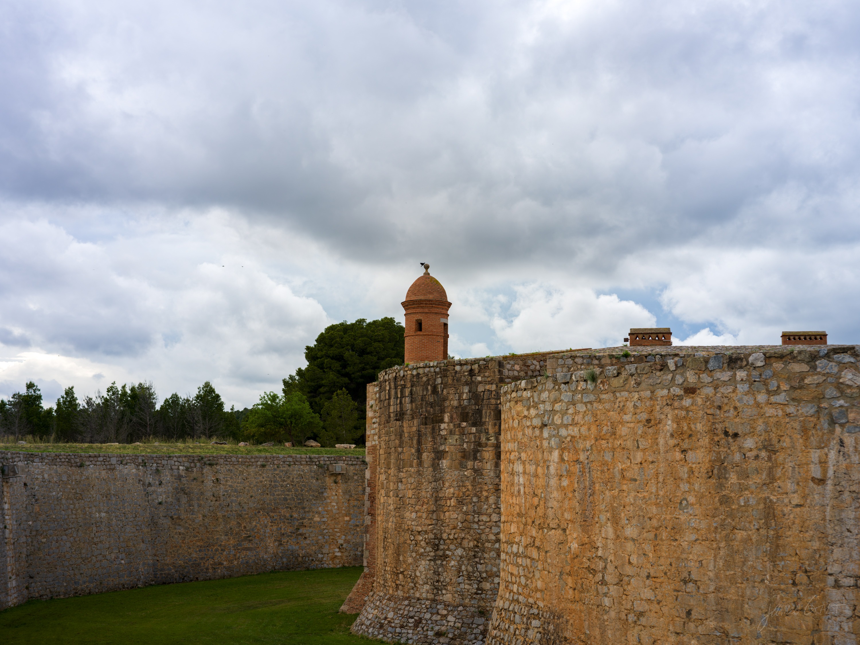 The outer wall of a fortress with a small round sentry turret on the corner, a dry green moat below and heavy cloud above.
