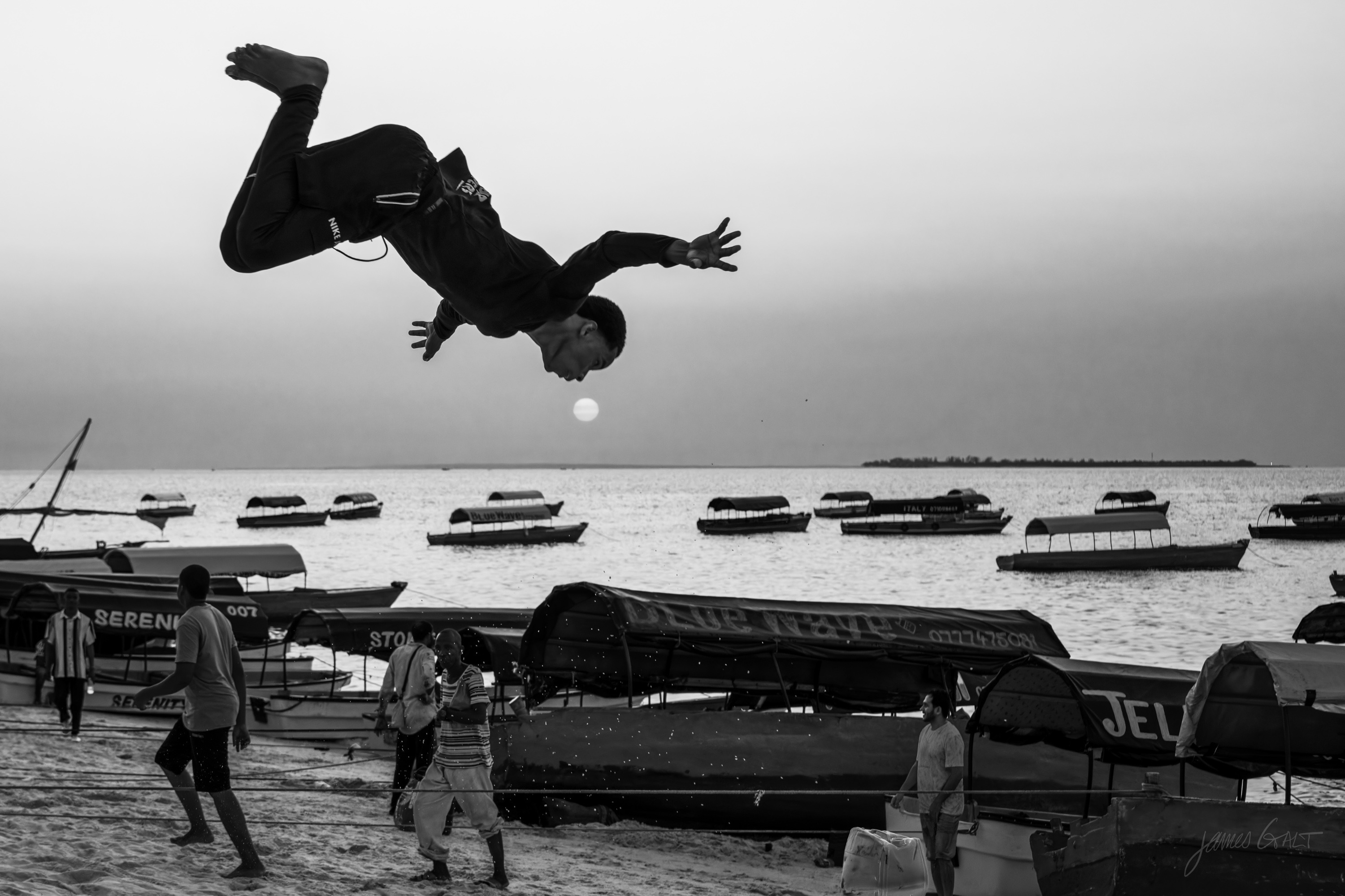 Black and white photograph of a young man inverted at the top of a somersault high over the beach, body curled, against a plain sky.