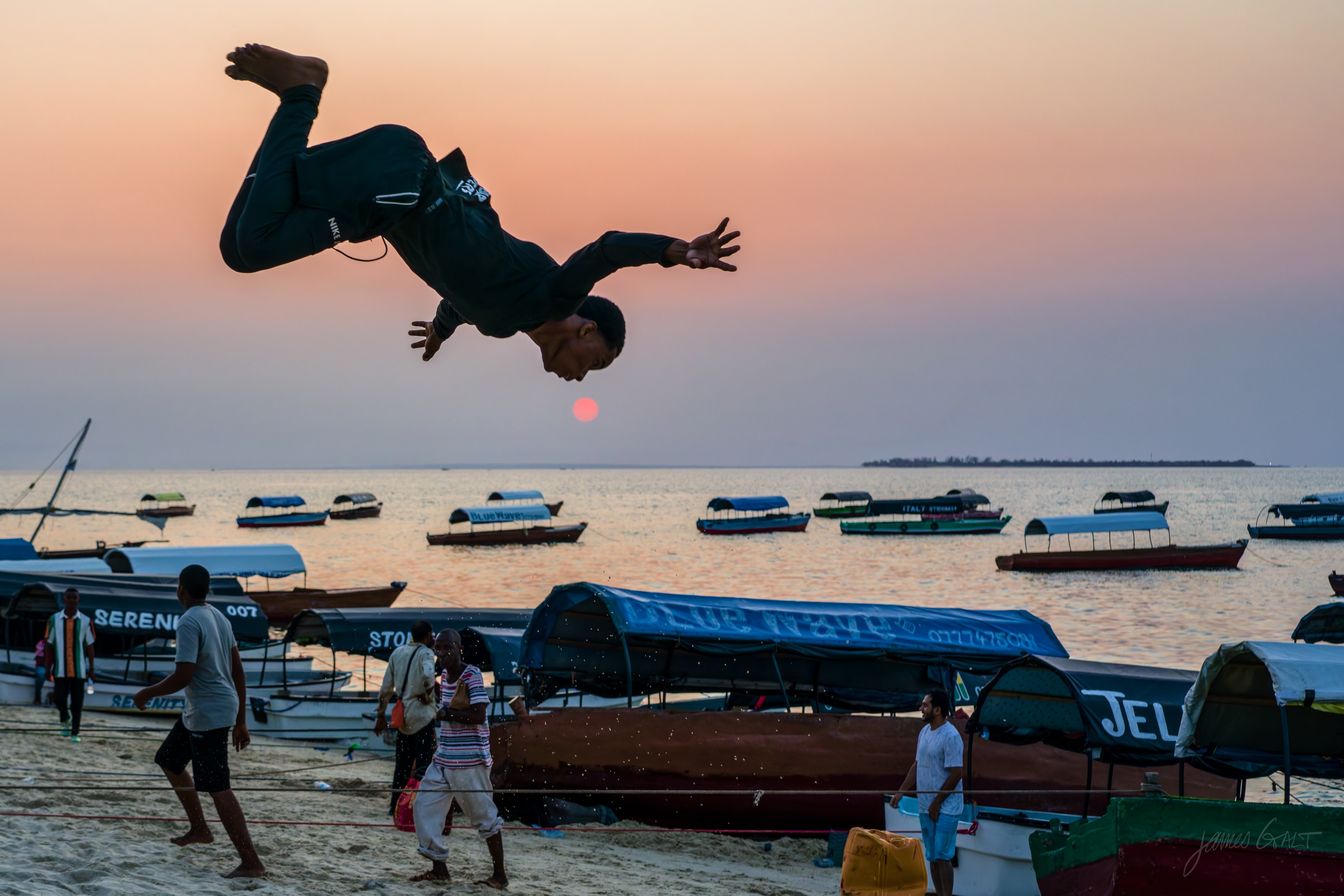 The same somersault in colour, the acrobat upside down in mid-air against a warm dusk sky, the beach and boats below.