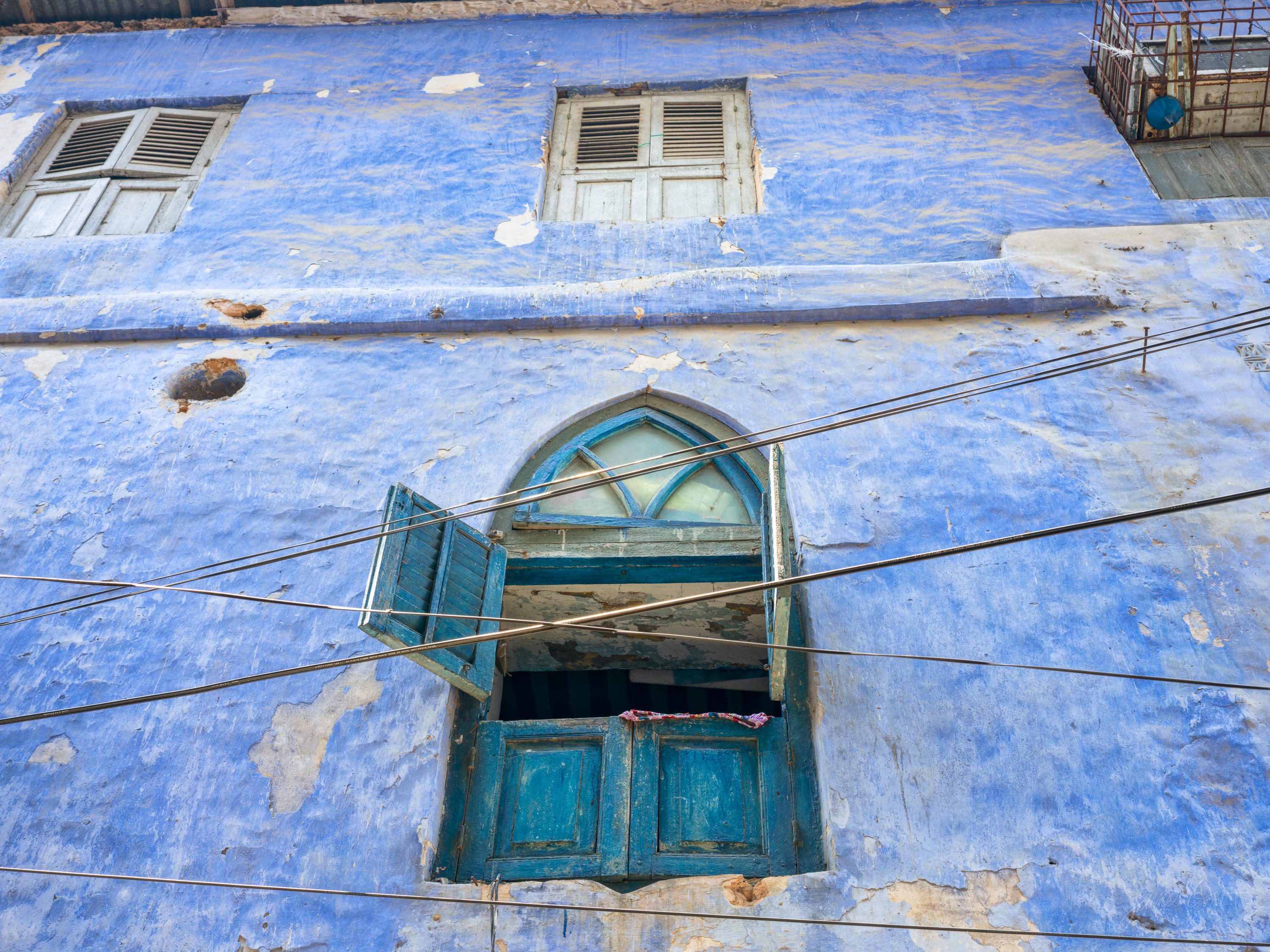 A weathered building painted deep blue with a carved wooden window and shutters set into the wall, the paint peeling at the edges.