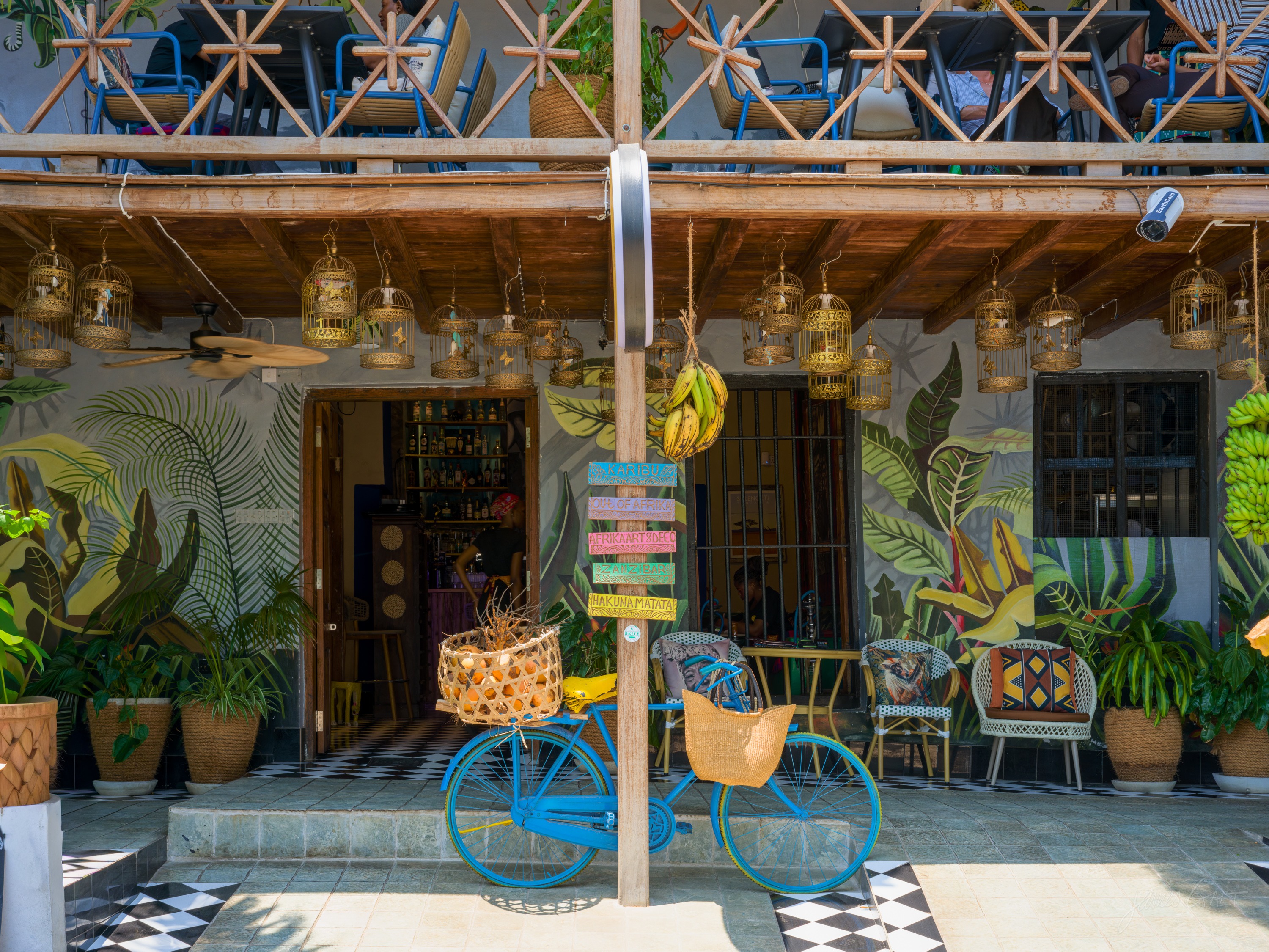 A brightly painted cafe front in Stone Town, colourful walls and hanging lamps, plants and chairs out on a tiled floor.