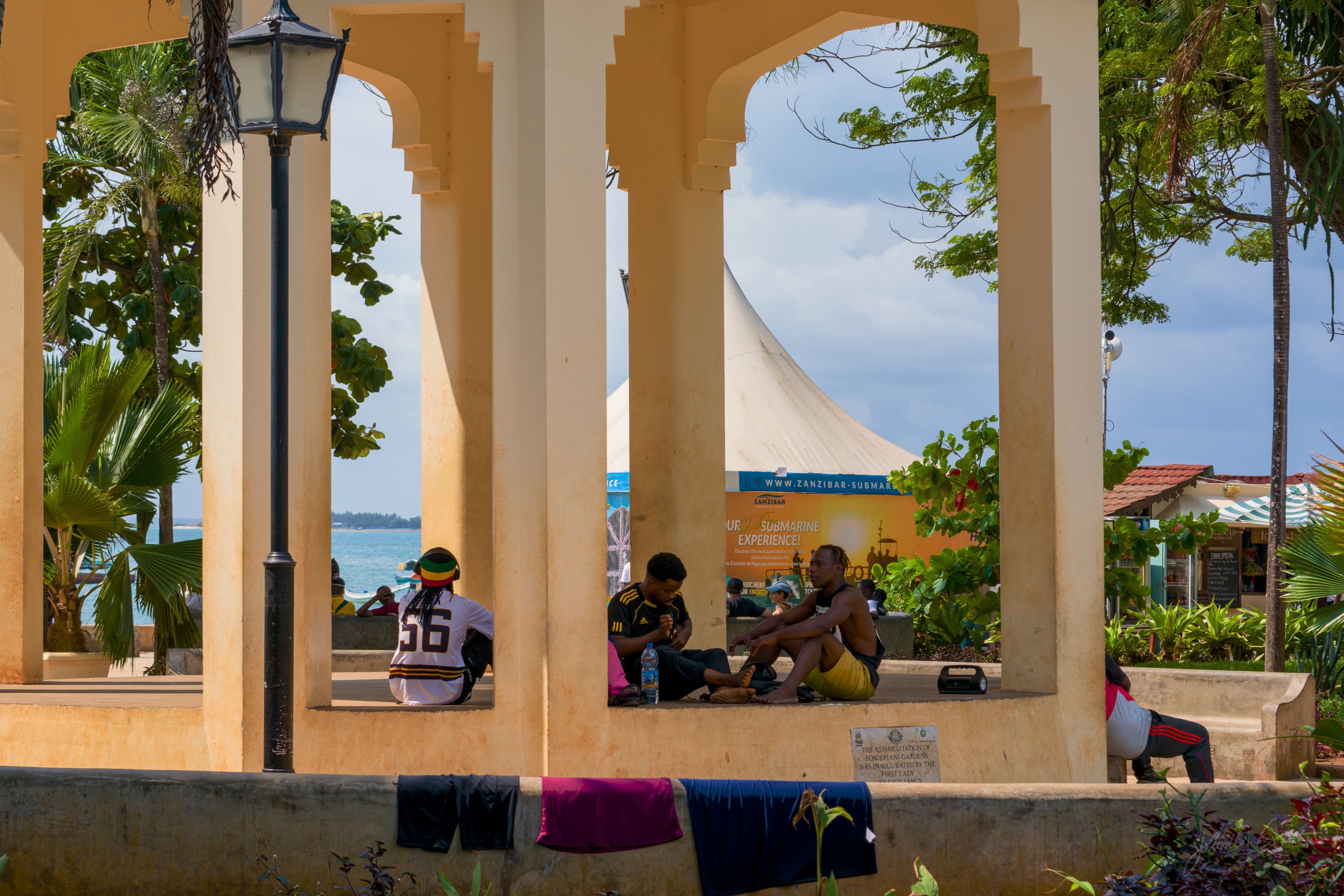 An arched stone colonnade along the seafront, the sea and a distant sail visible through the arches, people resting in the shade.
