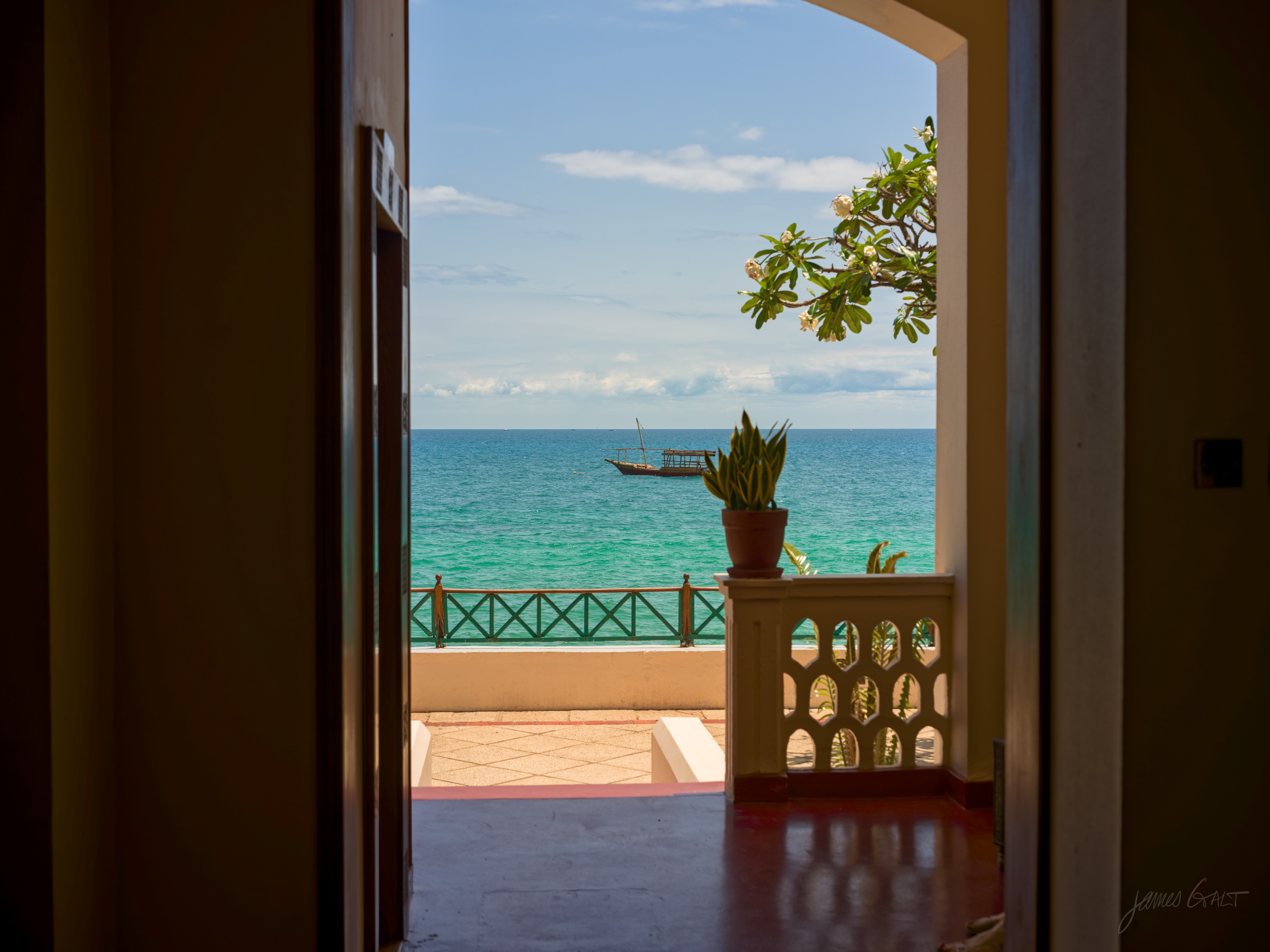 A dark interior doorway framing a bright view of turquoise sea and a carved balcony beyond, a plant silhouetted in the opening.