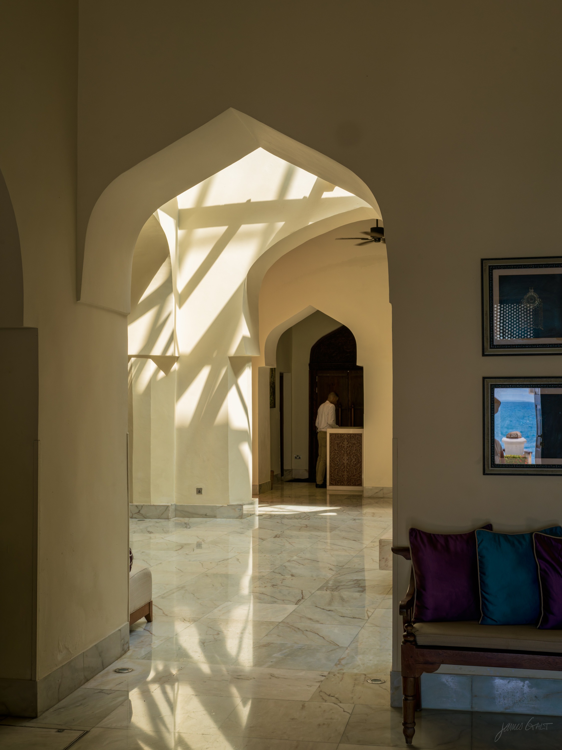A long arched hallway inside a Stone Town building, a series of pointed arches receding toward a bright doorway, cushions and pictures along the wall.