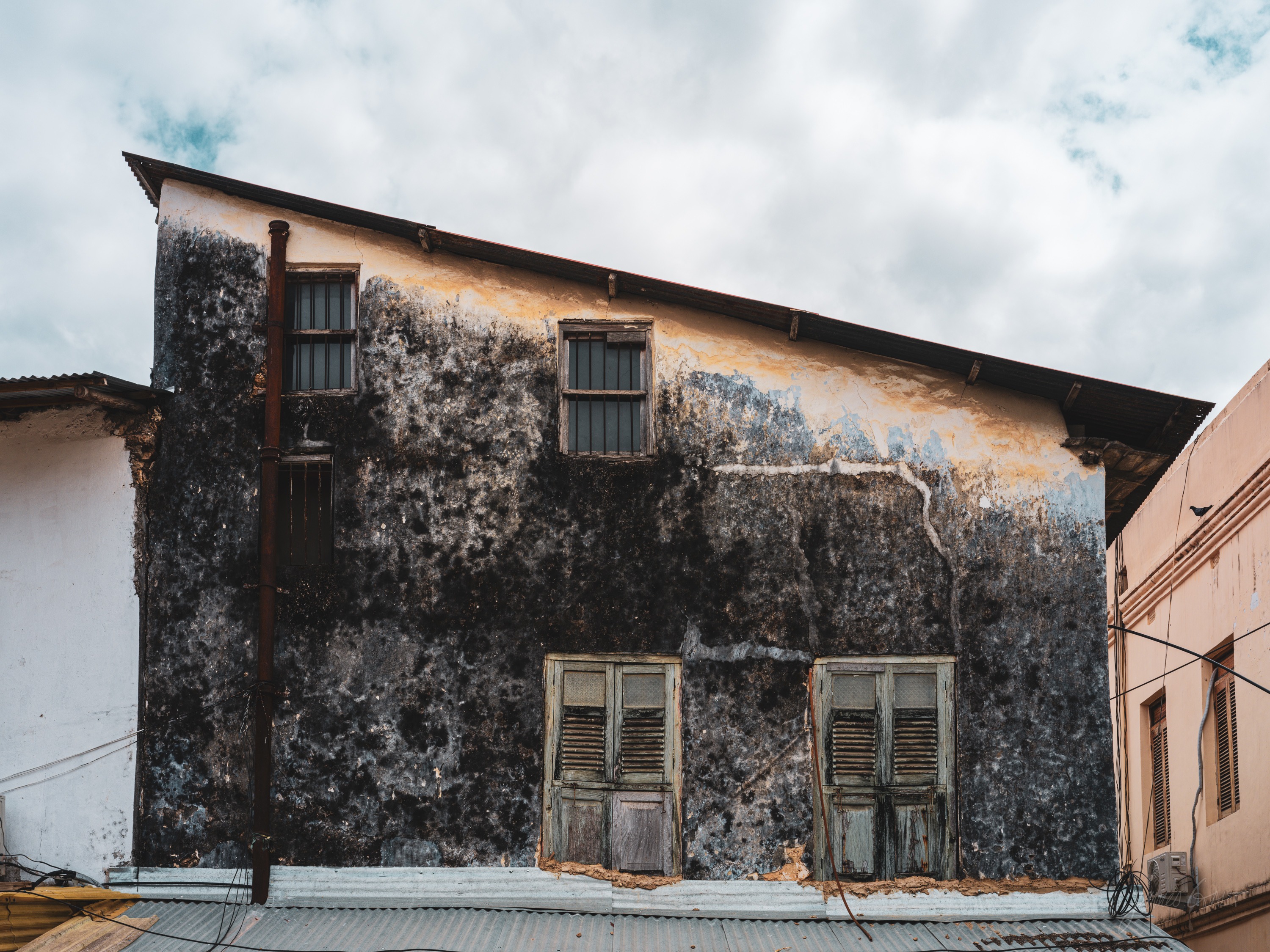 The facade of an old Stone Town building, plaster worn off in patches to the coral stone beneath, shuttered windows and a balcony, a pale cloudy sky above.