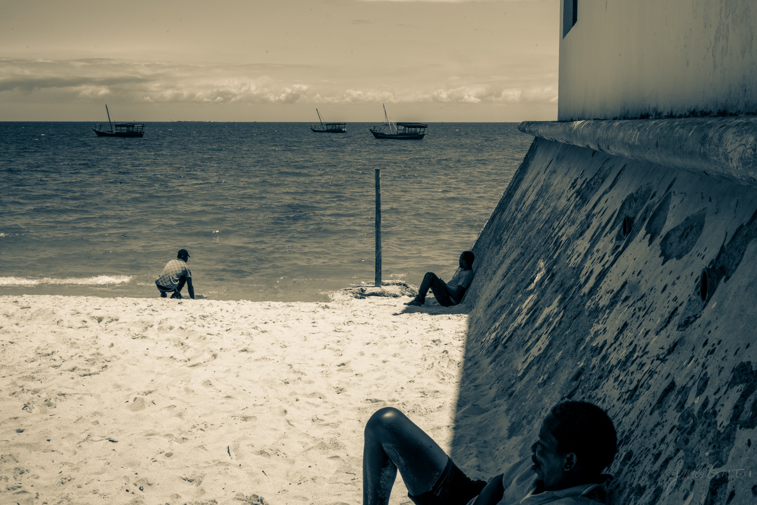 Black and white photograph of a pale beach below a curved stone sea wall, a lone figure sitting on the sand, boats out on the water beyond.