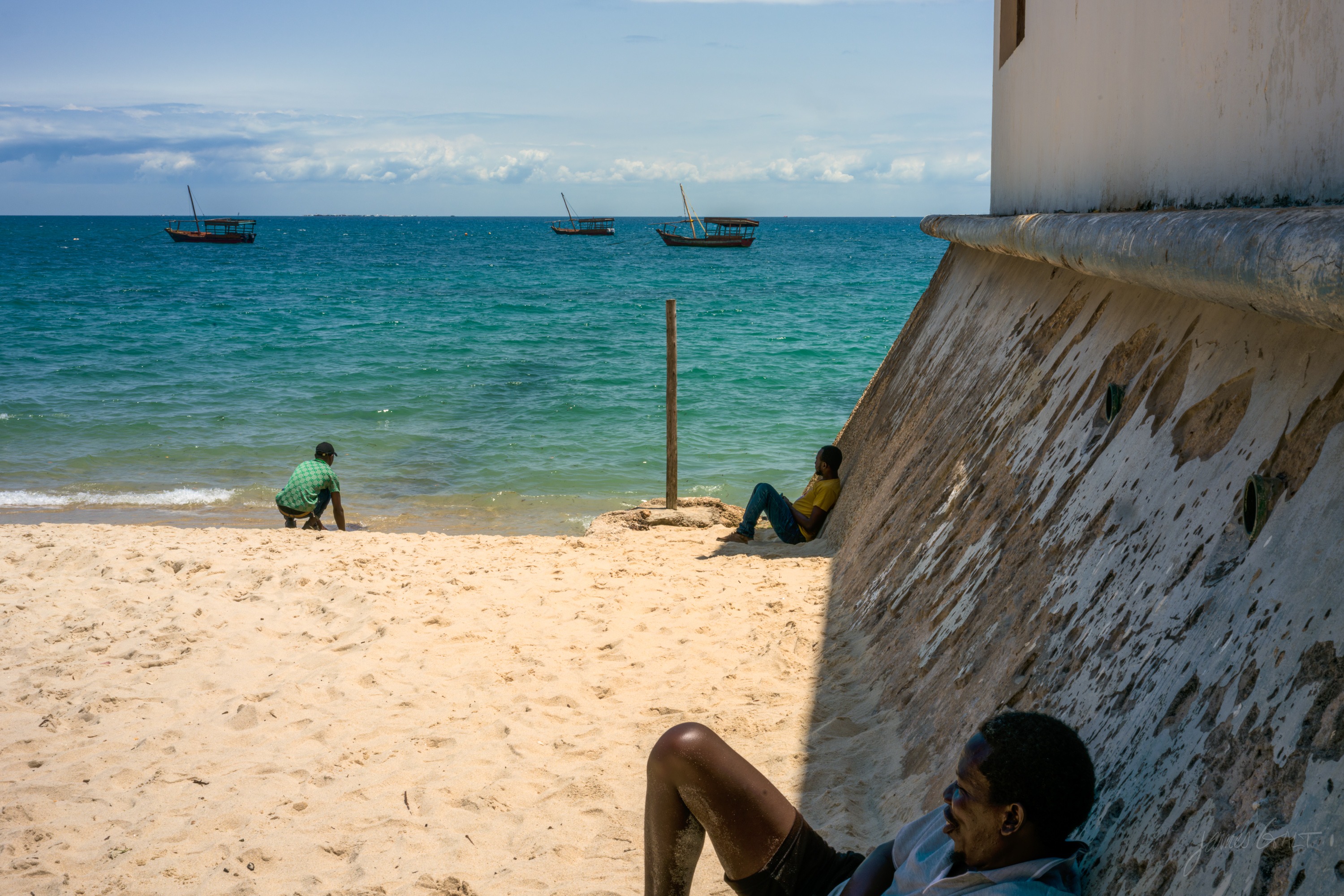 The same scene in colour, a sandy beach under a stone sea wall with a person sitting near it, moored boats and a hazy sky on the water.