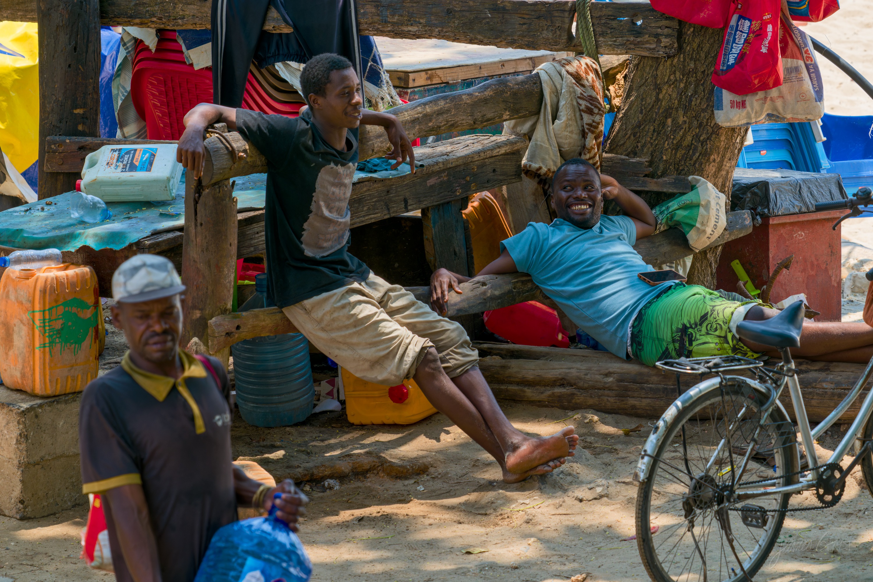 A group of men sitting and resting in the shade of a doorway and against a wall in a Stone Town lane, some talking, produce and a bicycle nearby.