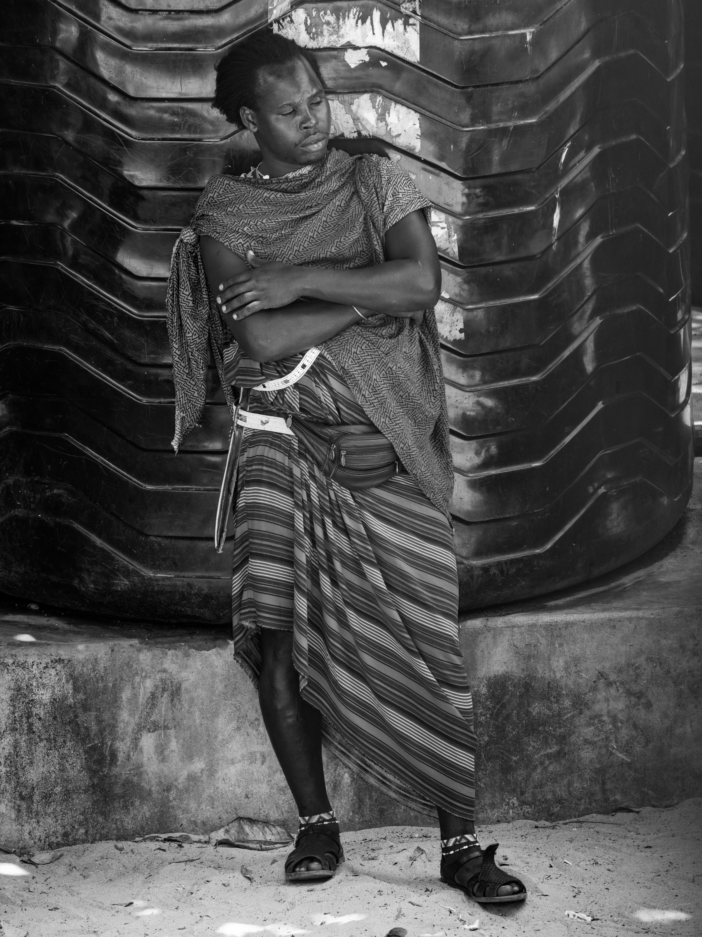 Black and white photograph of a Maasai man in a shuka leaning against a rough stone wall in a lane, standing in the shade.