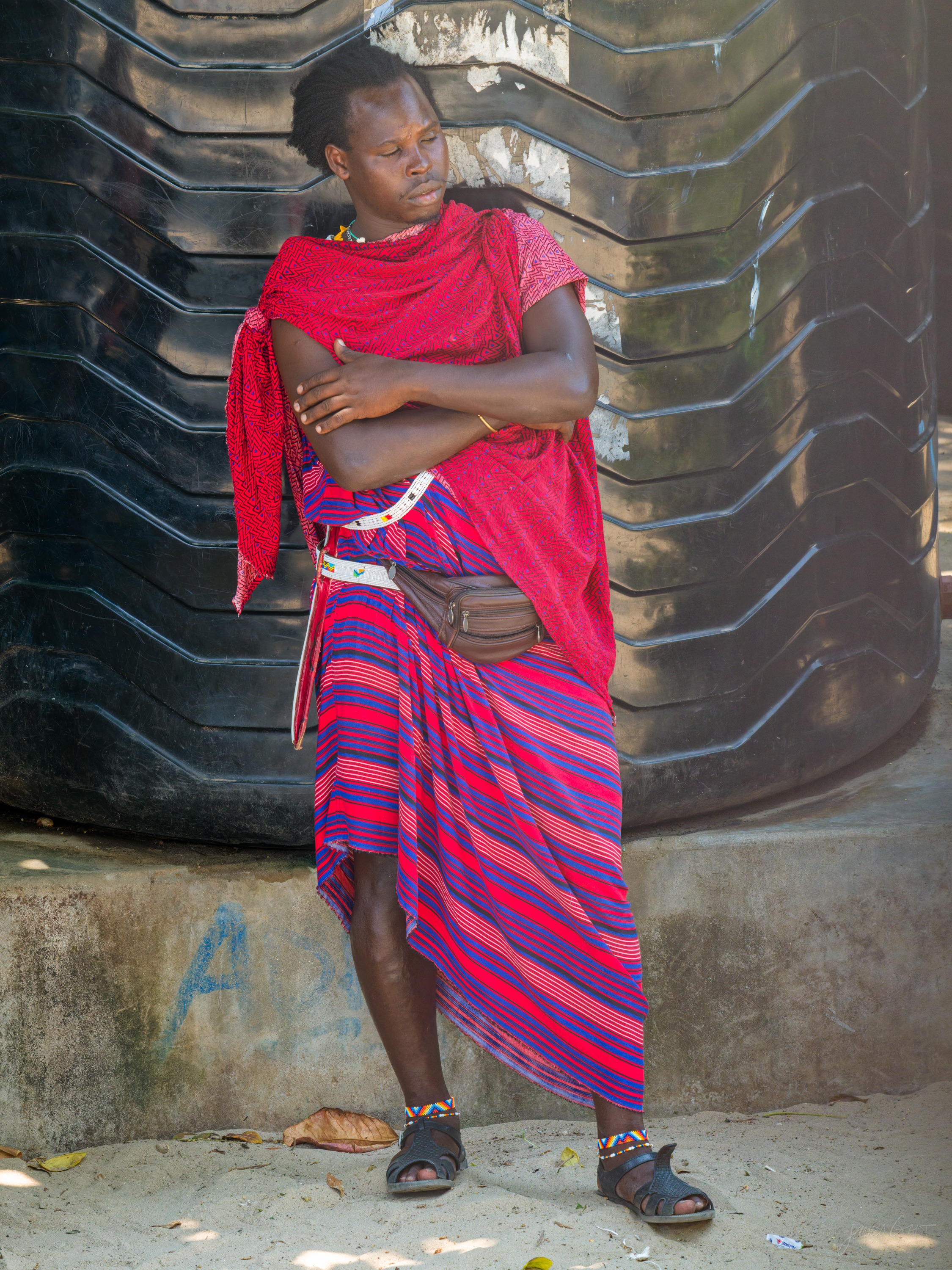 The same man in colour, in a red and pink checked shuka and beadwork, leaning against the weathered wall, sandals on his feet.