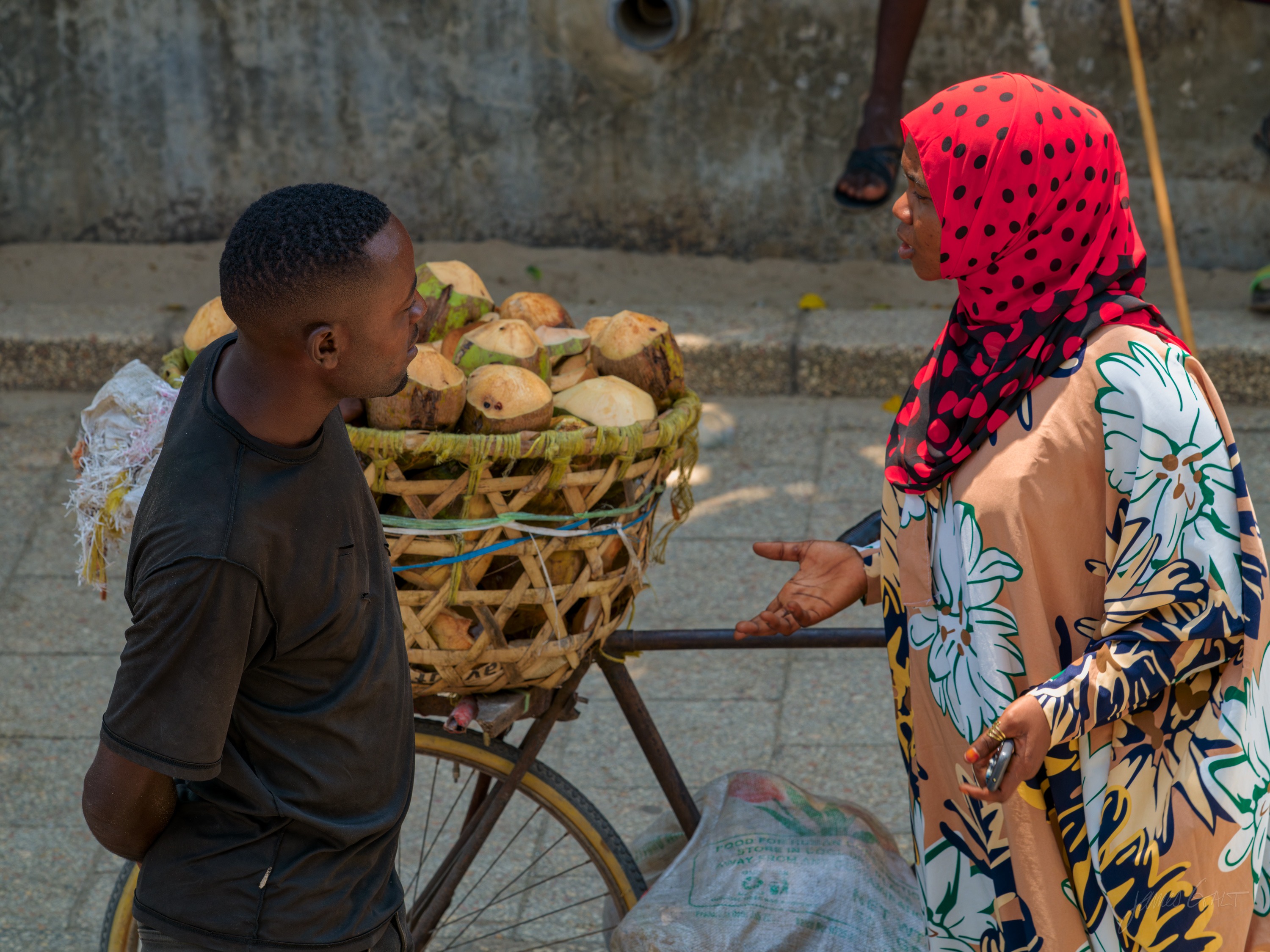 A man at a wooden fruit cart stacked with round fruit talking to a woman in a red headscarf, a Stone Town lane behind them.