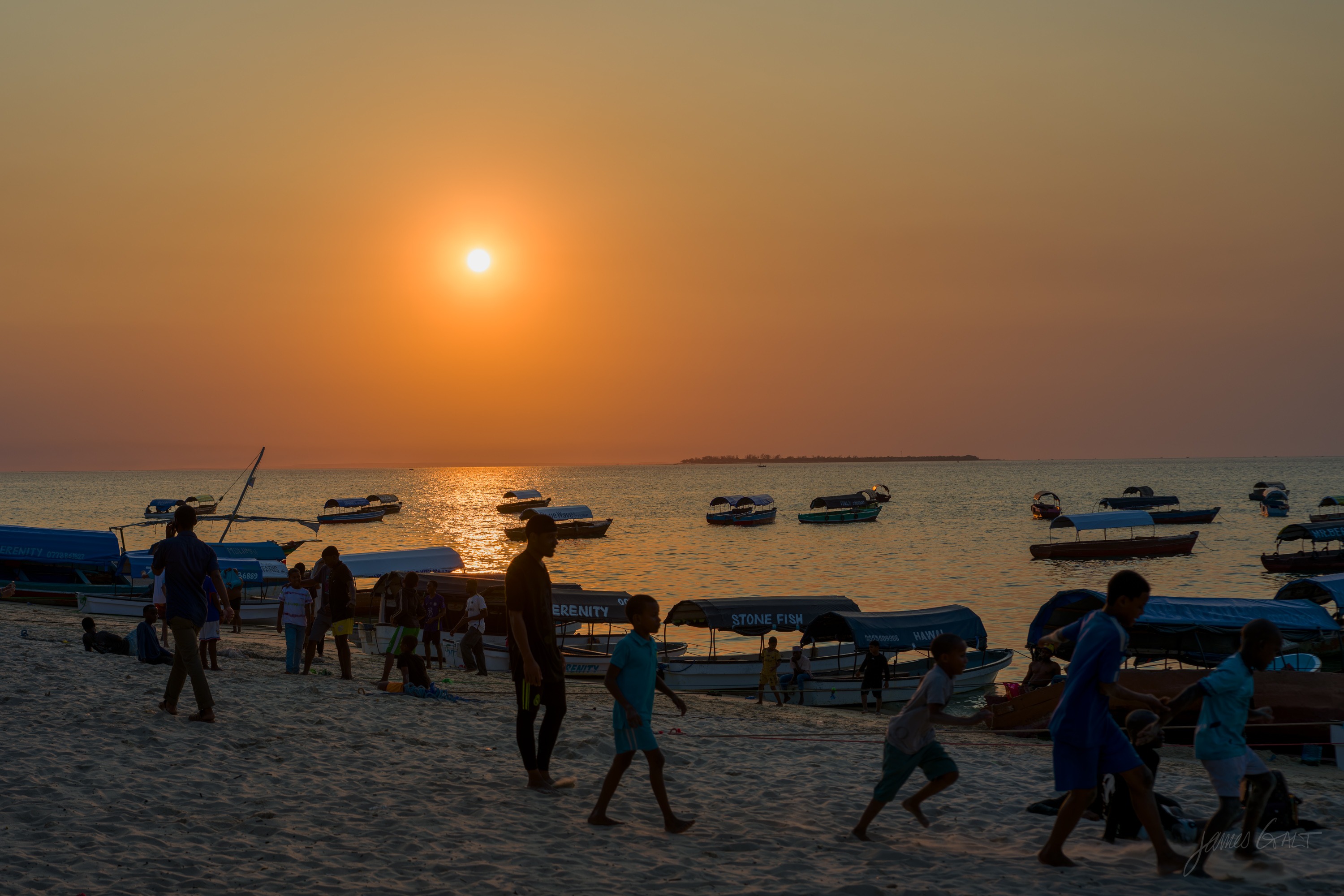 A hazy sunset over the Stone Town harbour, the low sun above a line of wooden boats, small figures on the beach in silhouette.
