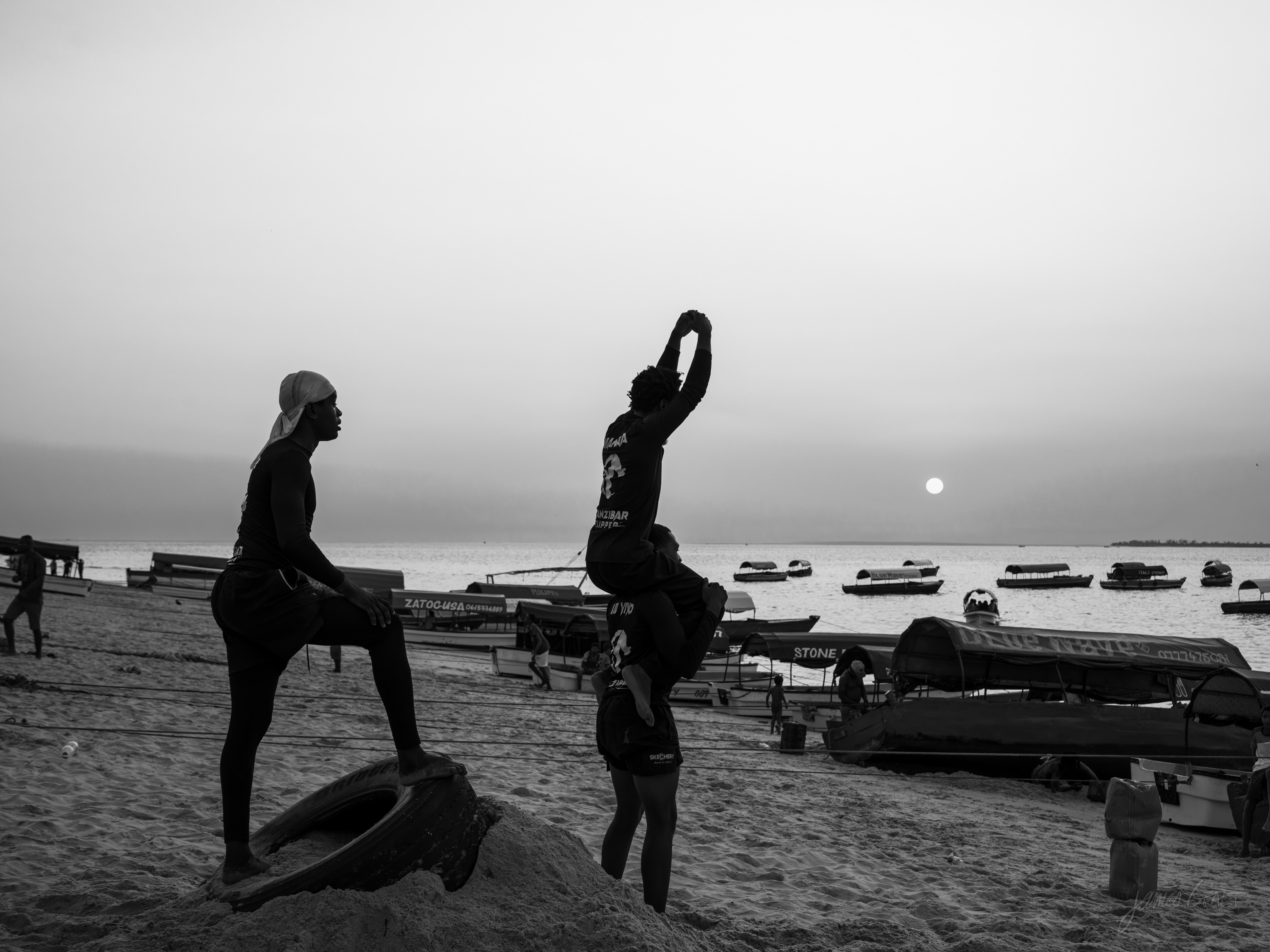Black and white photograph of a young man mid-somersault high above the beach at sunset, body tucked, boats and figures below.