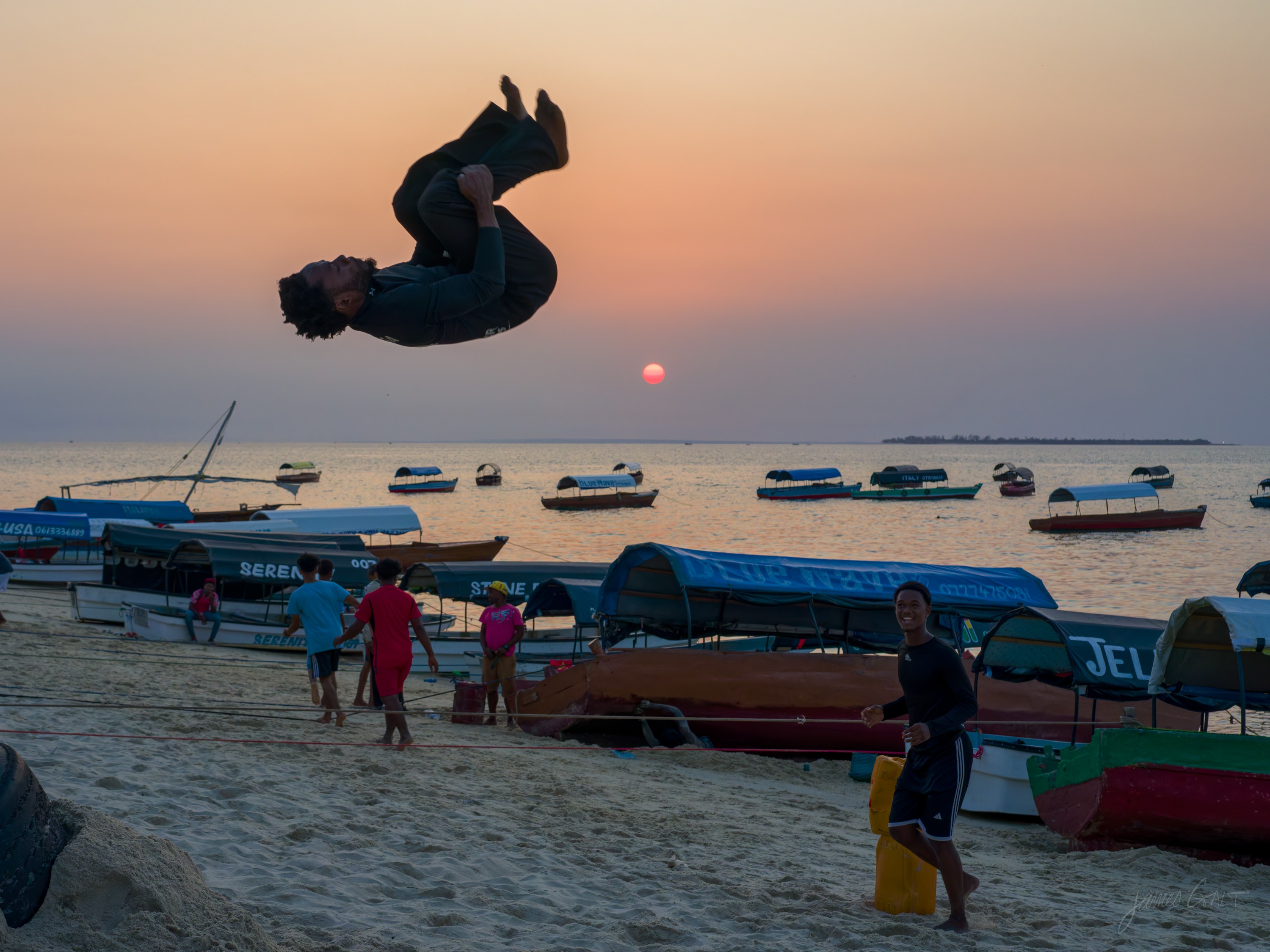 A young man leaping and flipping on the beach at sunset in colour, the warm sky and moored dhows behind, other people watching.