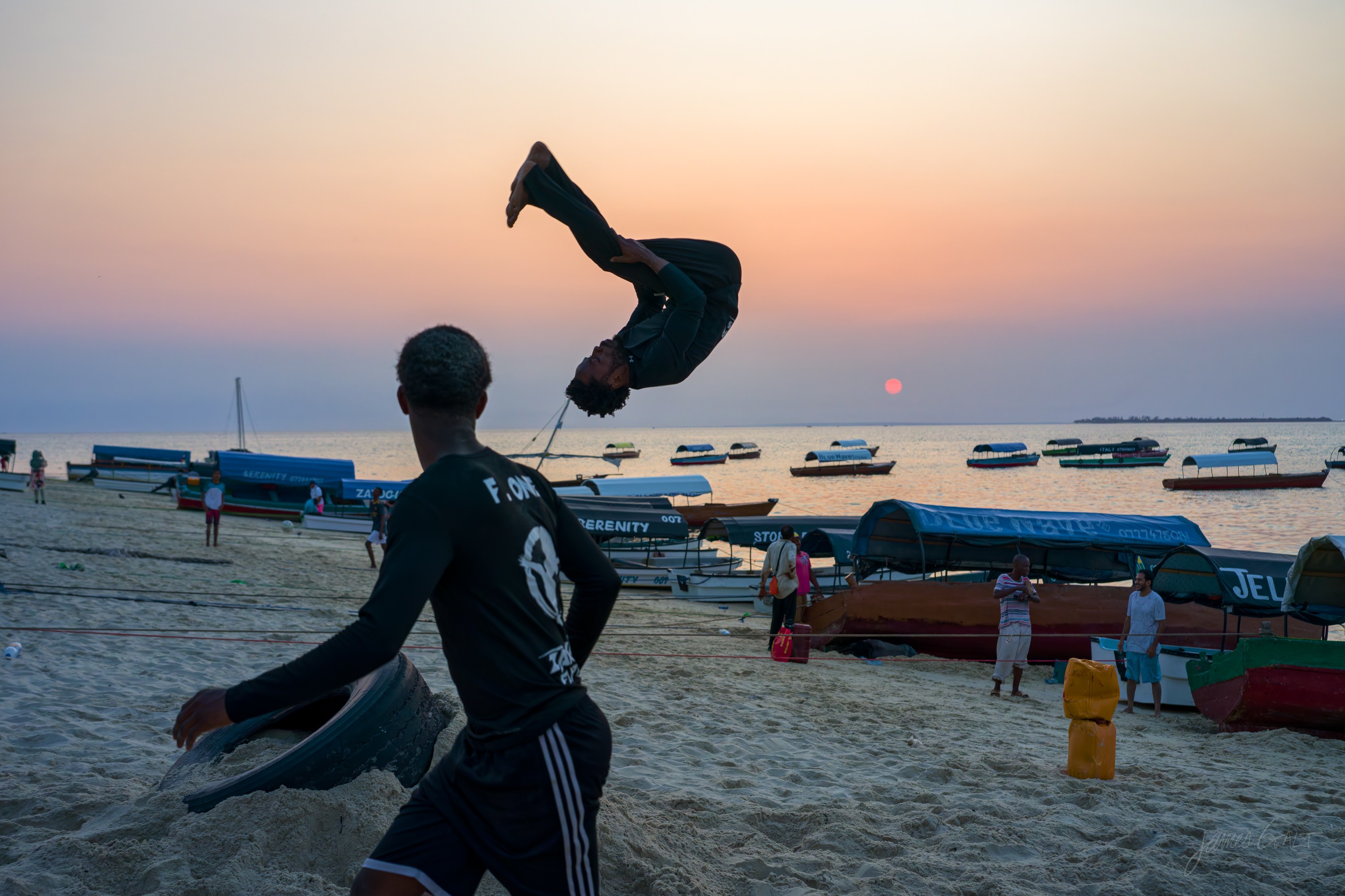Another acrobat mid-air over the sand at dusk, arms out, the sea and boats and a soft orange sky behind.