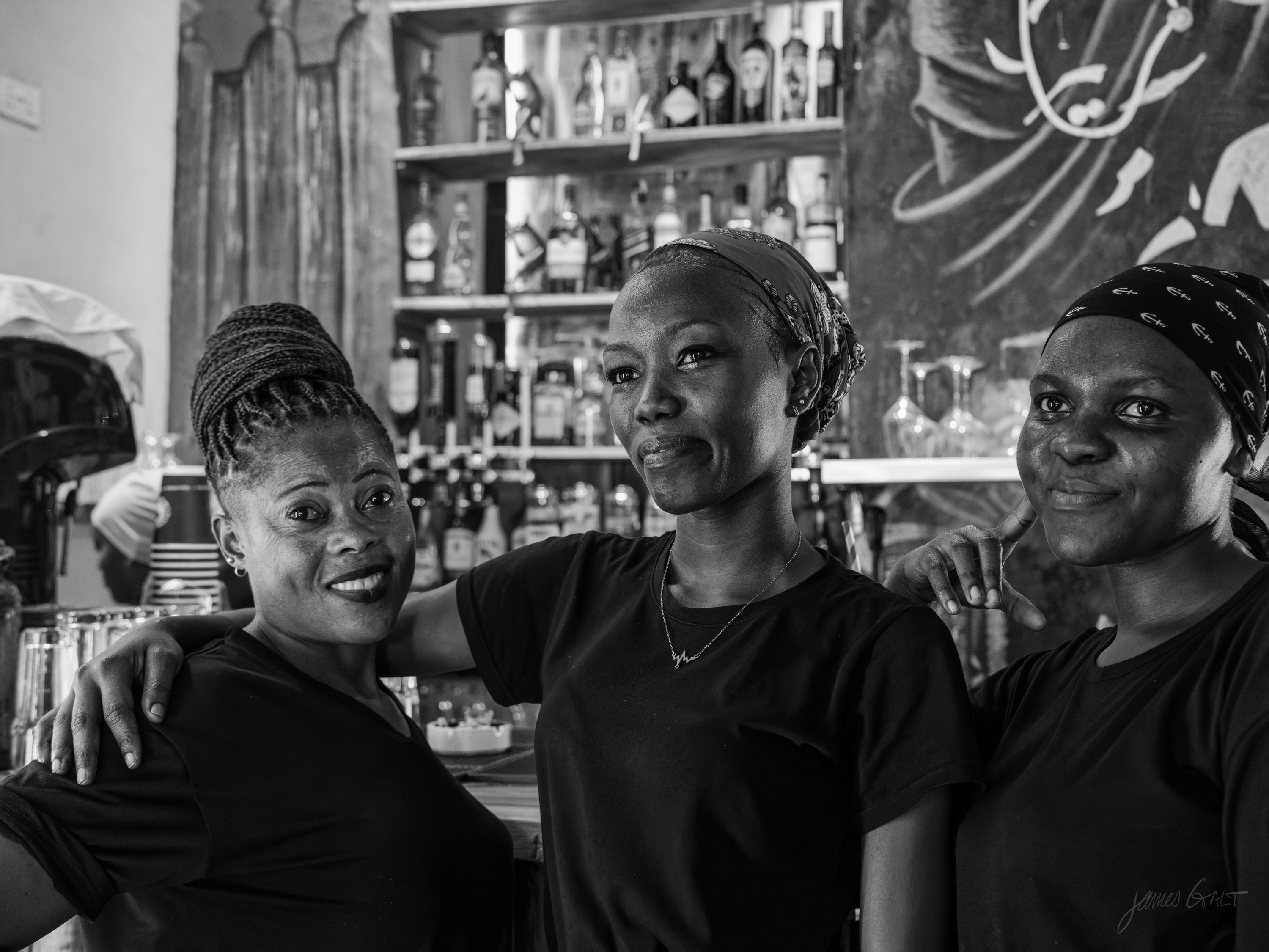Black and white photograph of three women behind a bar, one with an arm around another, all mid-laugh, bottles and a chalkboard behind them.