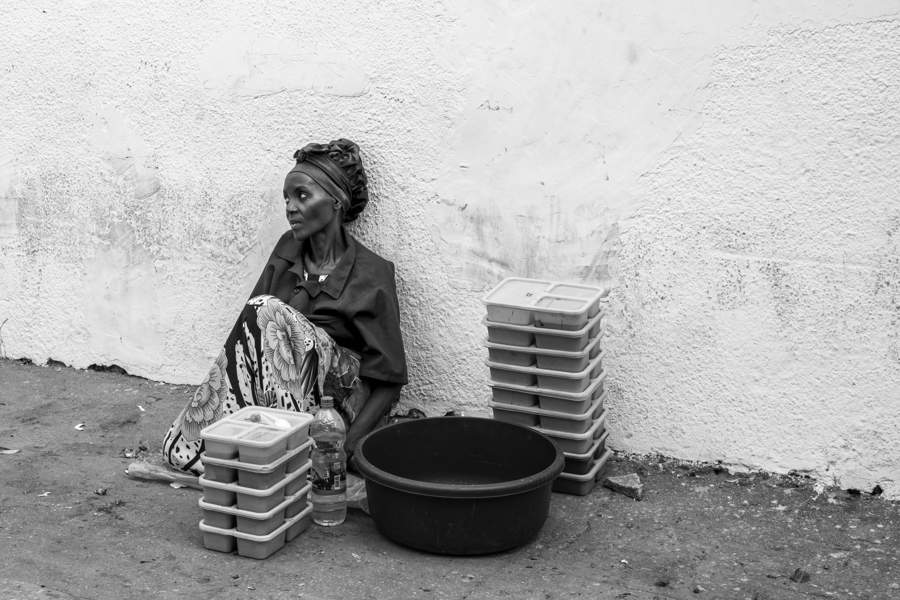 Black and white photograph of a woman sitting on the ground against a white wall selling stacks of plastic food containers, a basin beside her, tightly framed.