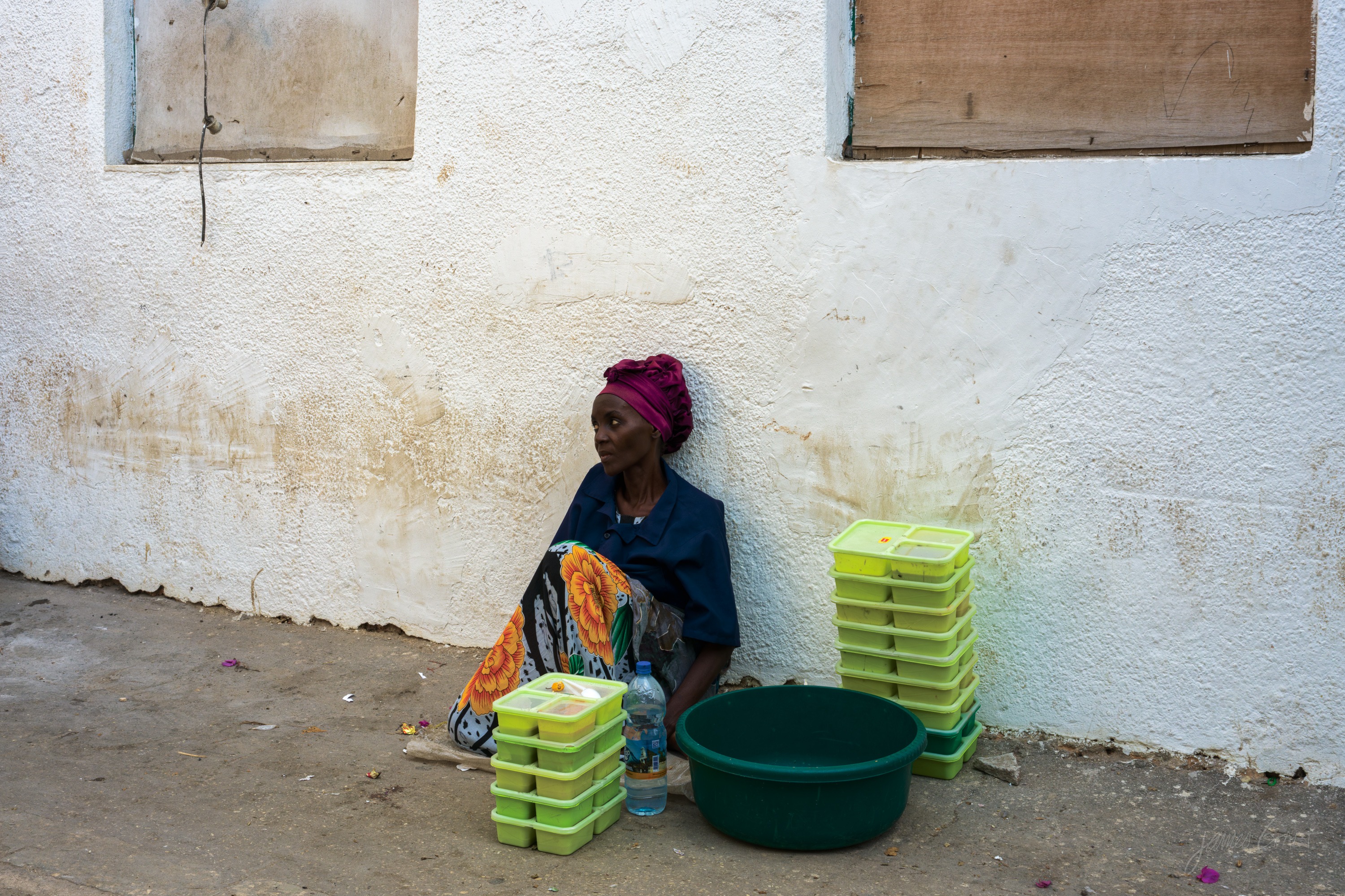 The same scene in colour, the woman in patterned cloth on the ground with green and pink containers stacked around her against the pale wall.