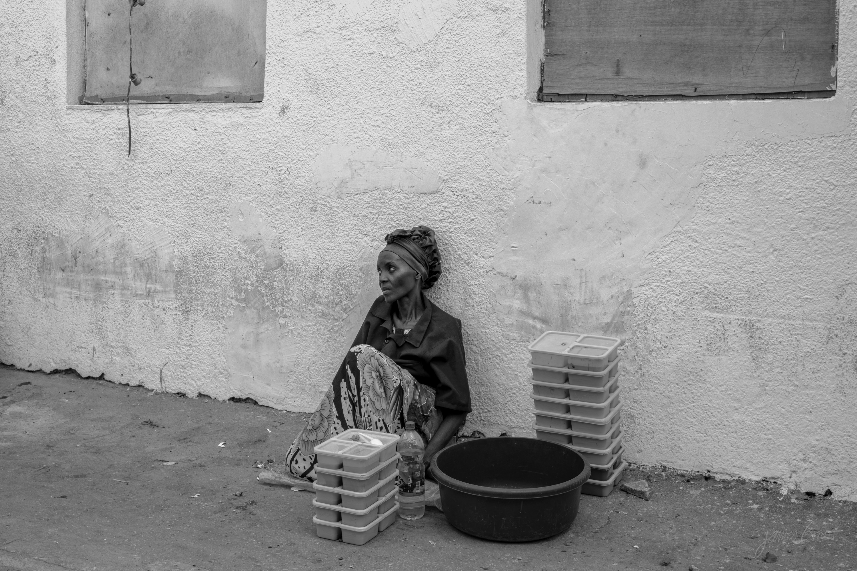 A wider black and white view of the woman against the wall, two shuttered windows above her, the stacks of containers and basin around her.