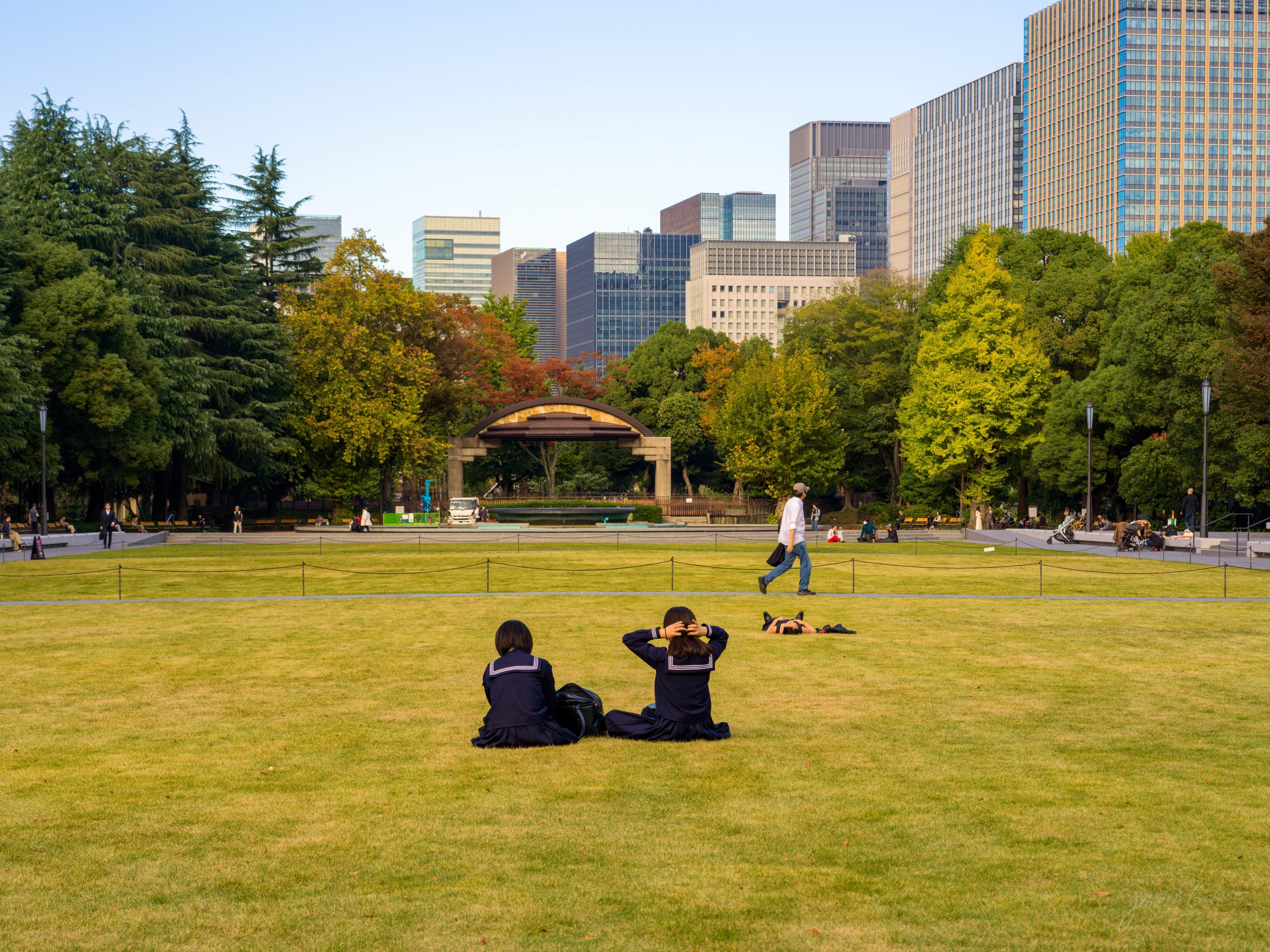 A broad sunlit park lawn with two children in dark school uniforms sitting on the grass, their backs to the camera. Beyond a low fence sit a domed open-air bandstand, autumn trees, and a wall of glass office towers. A person walks across the middle distance and another lies on the grass.