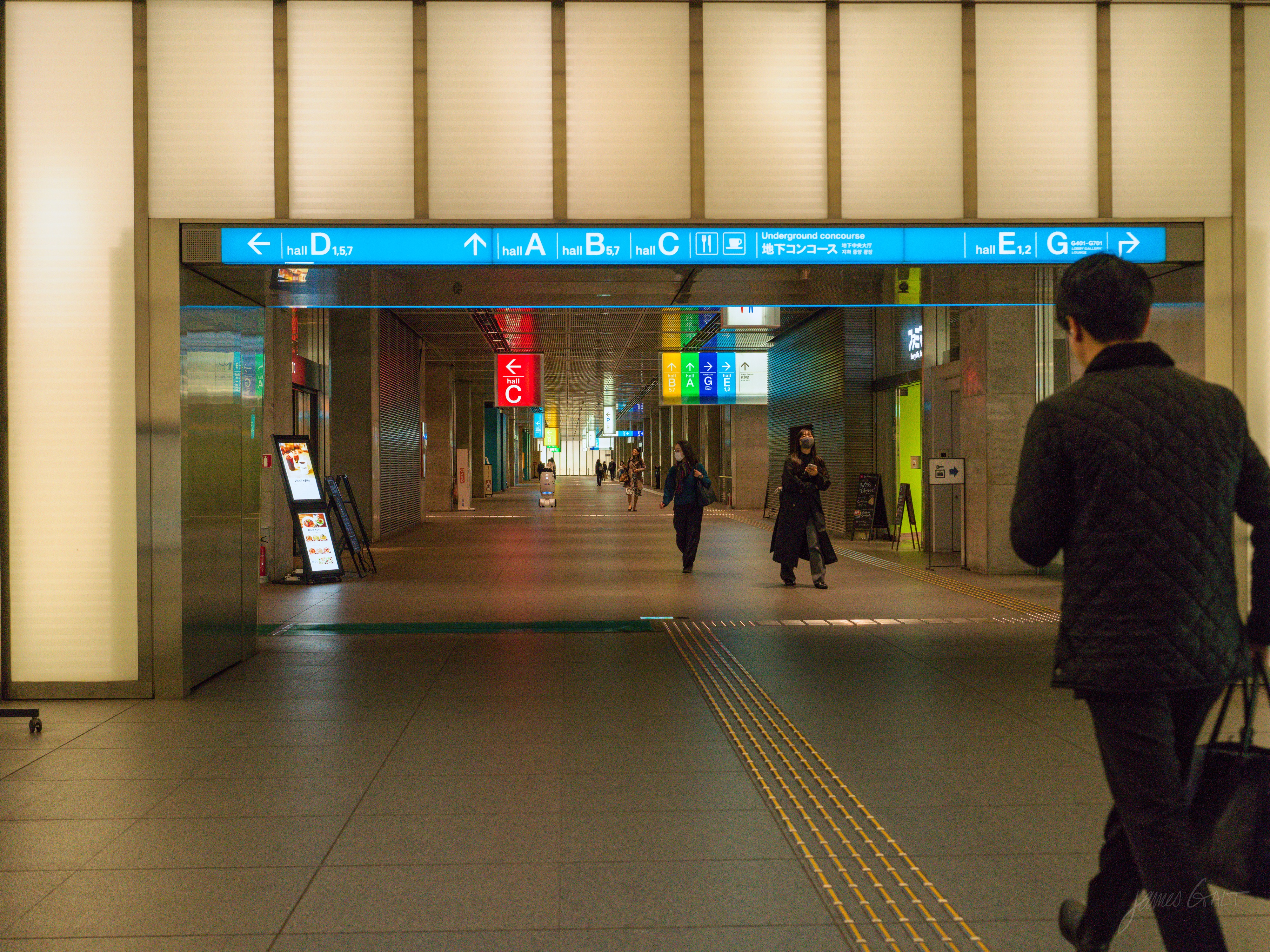 An underground station concourse lit warm and dim, with overhead blue signage pointing to exits A through G. A man in a quilted jacket walks toward the right; two other figures stand further down the passage, with tactile paving running across the polished floor.