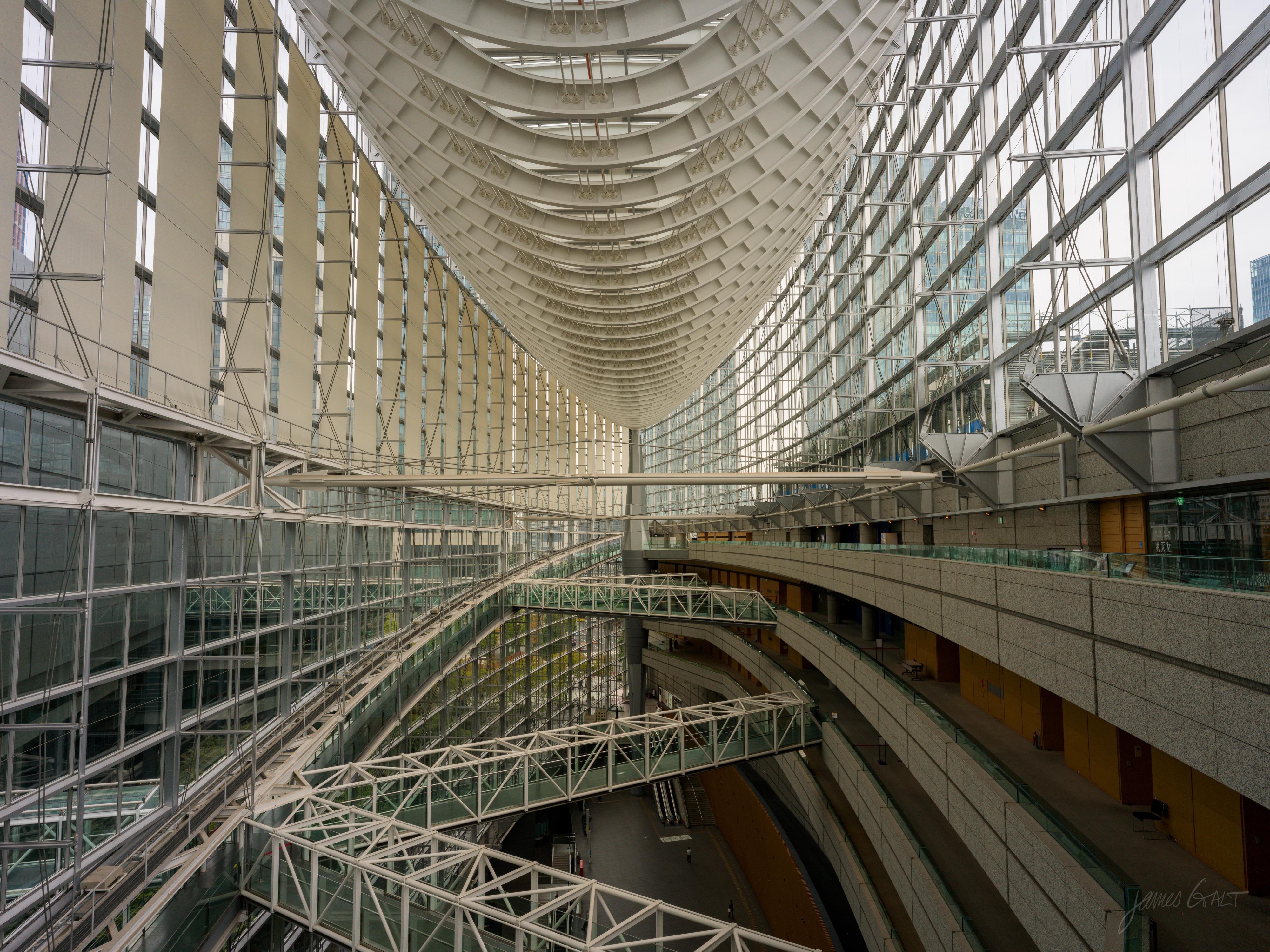 The interior of the Tokyo International Forum seen along its length, the boat-hull glass roof curving overhead and tapering into the distance, with steel walkways and bridges crossing the tall atrium below.