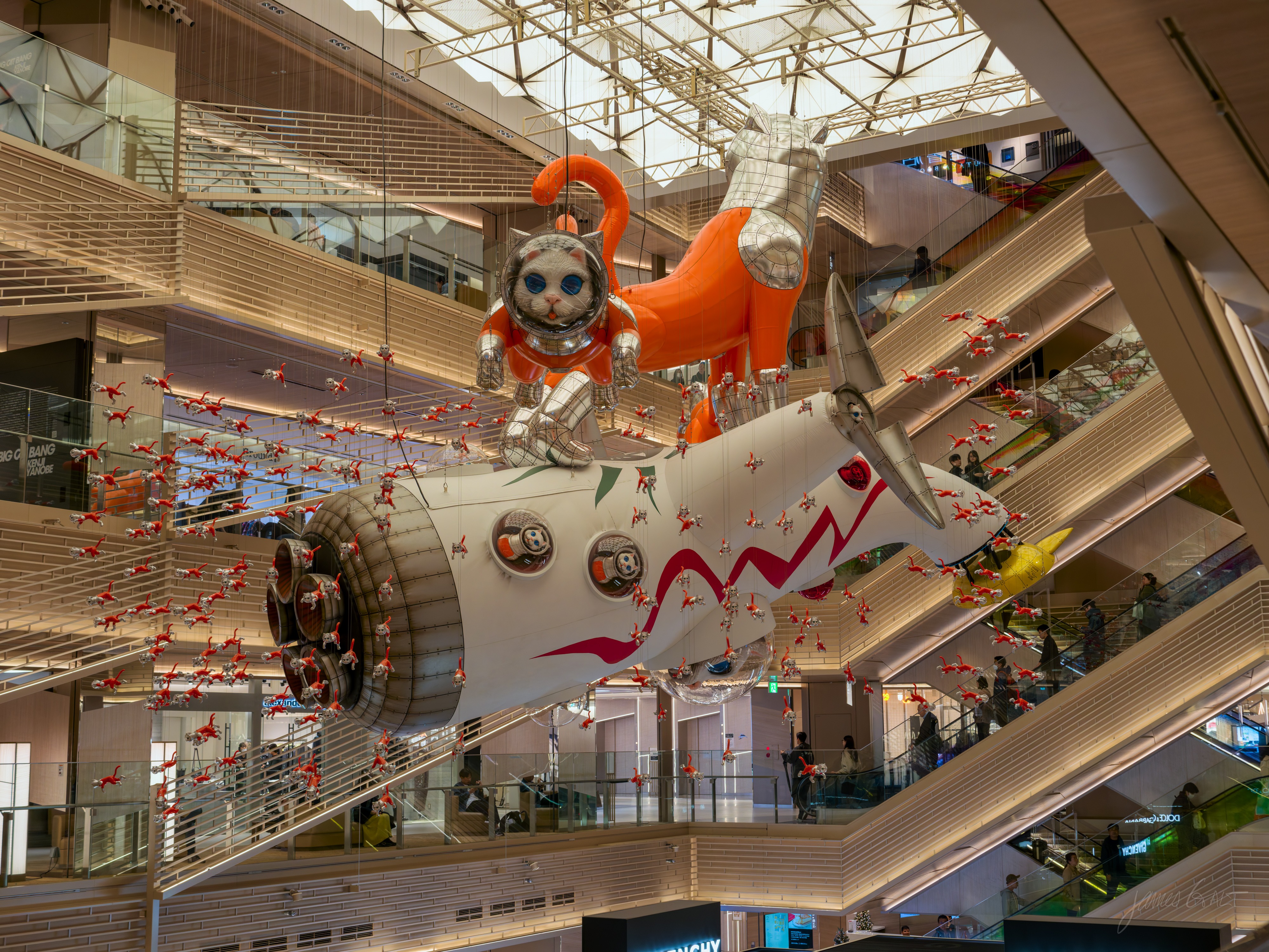 Looking up into a multi-storey shopping atrium at a large suspended sculpture: an orange cat in an astronaut suit riding atop a silver-and-white rocket shaped like a plane, surrounded by hundreds of small red darts strung on wires. Escalators and balconies recede on both sides.