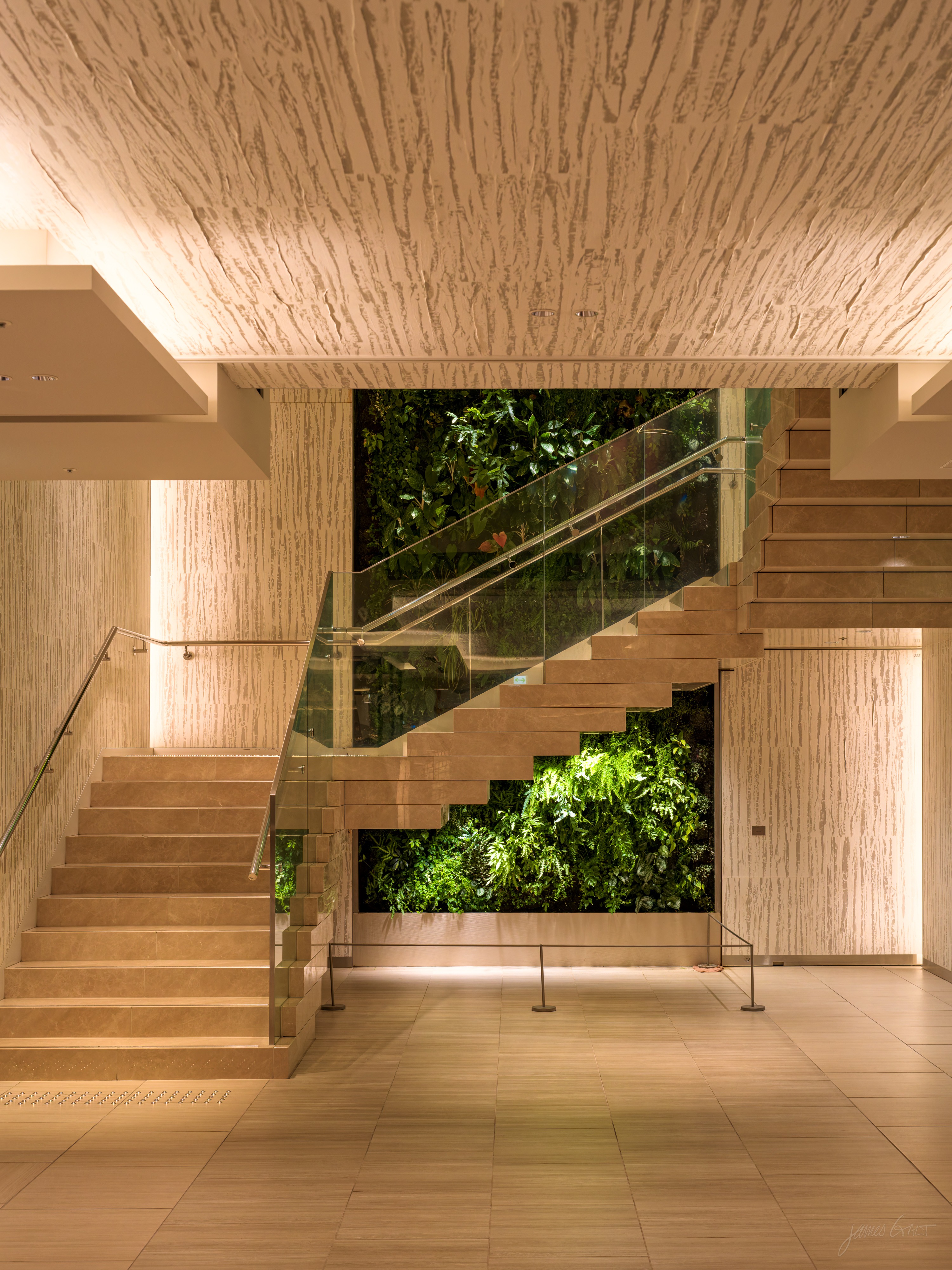 A warmly lit interior staircase faced in pale travertine, with a glass balustrade. Through the open stairwell, a two-storey wall of green foliage is visible. The ceiling has a heavily textured stone surface lit from below.